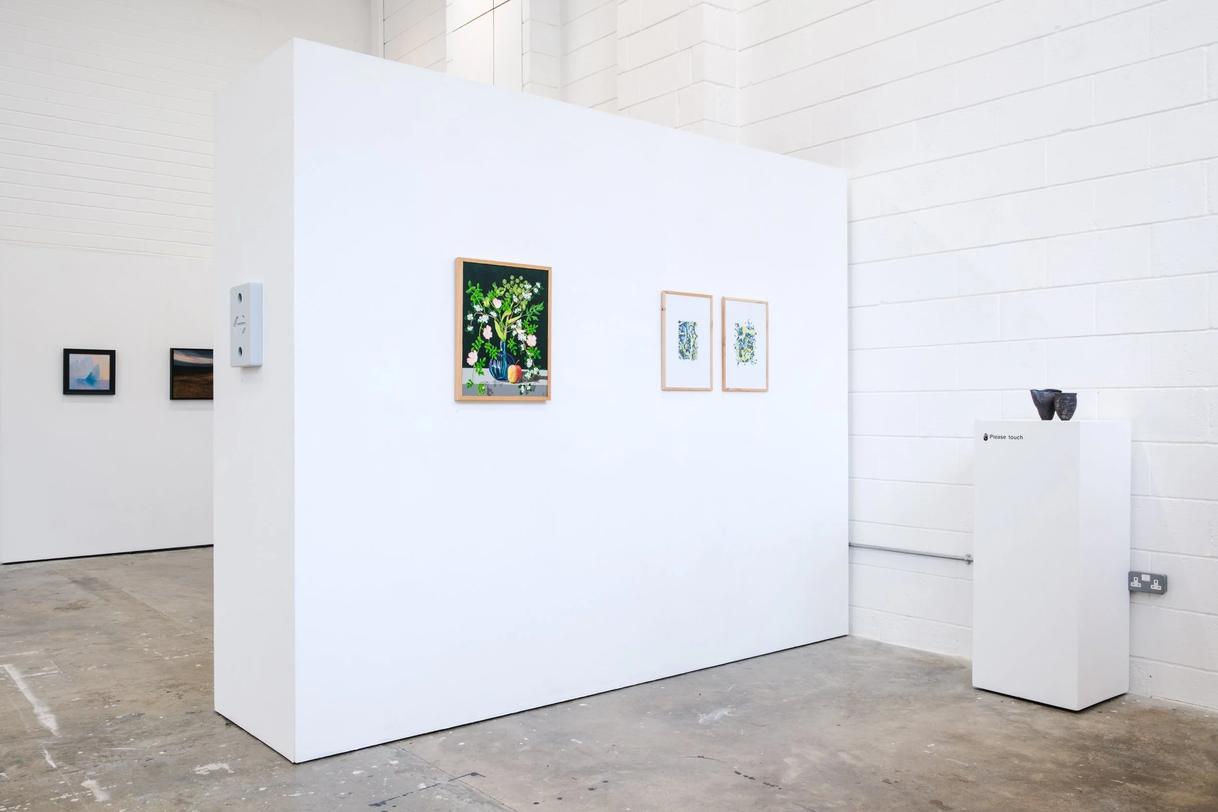Art gallery with framed pictures on white walls, small sculptures on a pedestal, and a white partition in the center of the space.