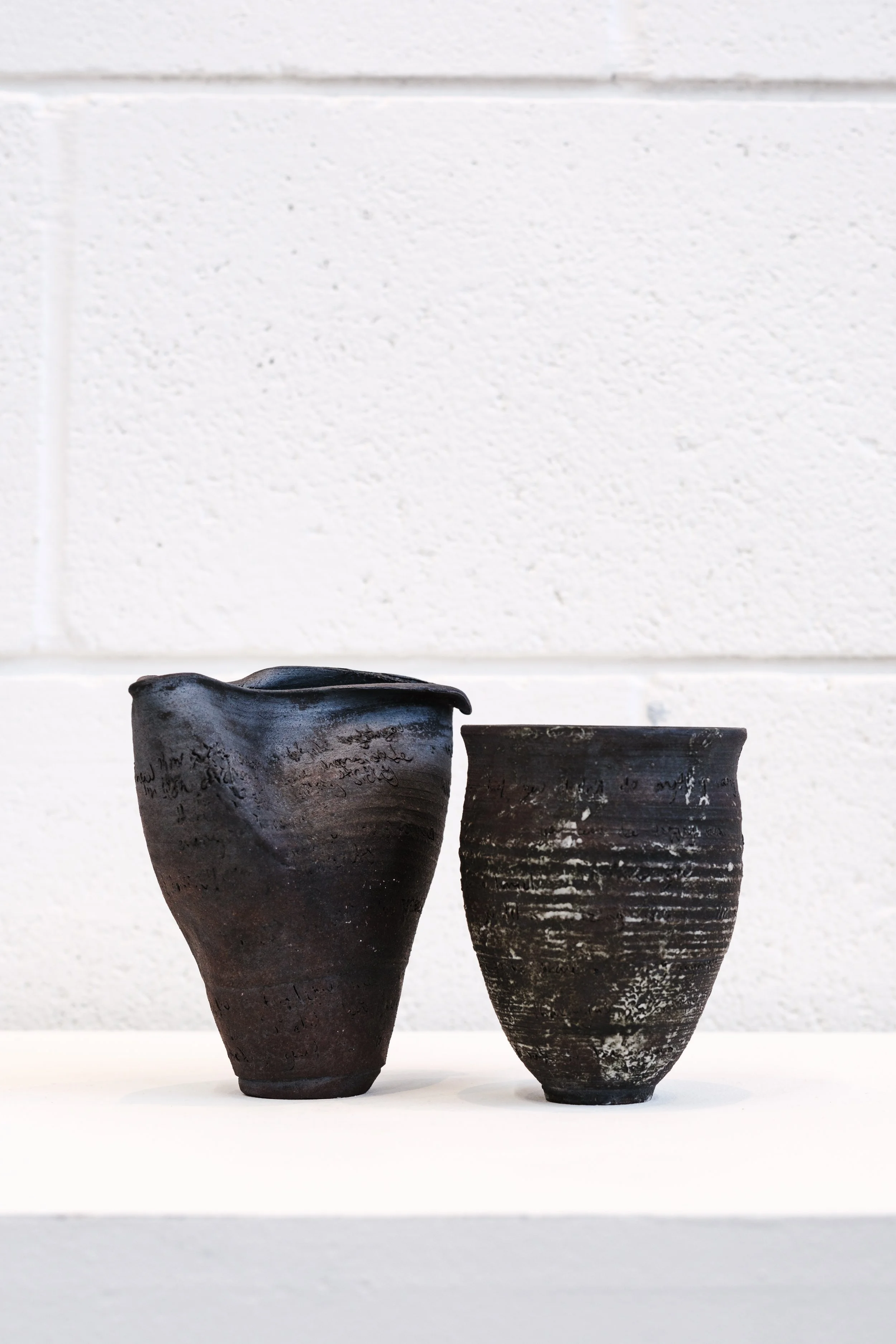 Two black ceramic vessels, one with a tall, rounded shape and the other shorter and wider, are placed on a white surface against a white brick wall background.