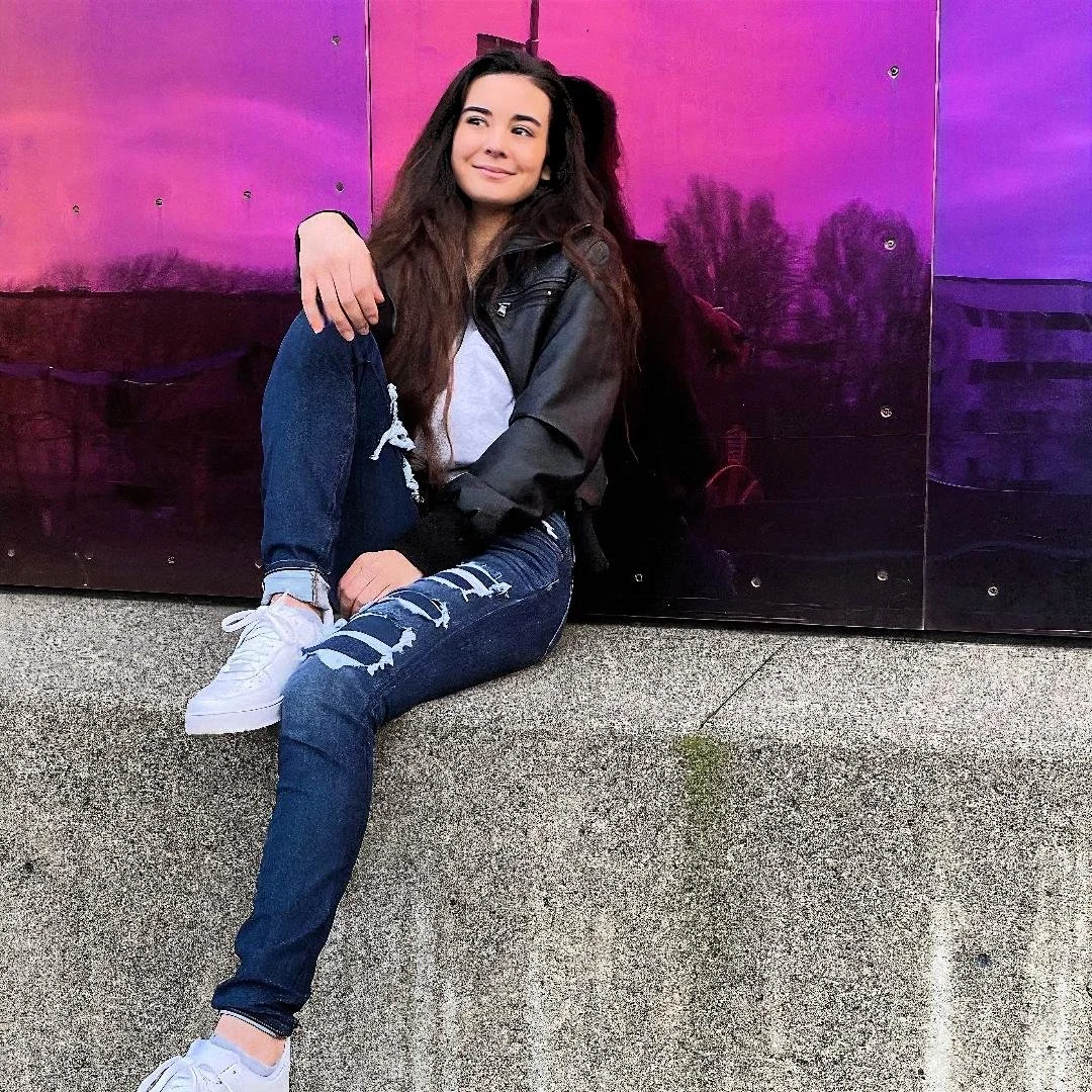 A young woman with long brown hair, wearing a black jacket, white t-shirt, ripped jeans, and white sneakers, sitting on a concrete ledge in front of a purple and pink reflective wall, smiling softly.