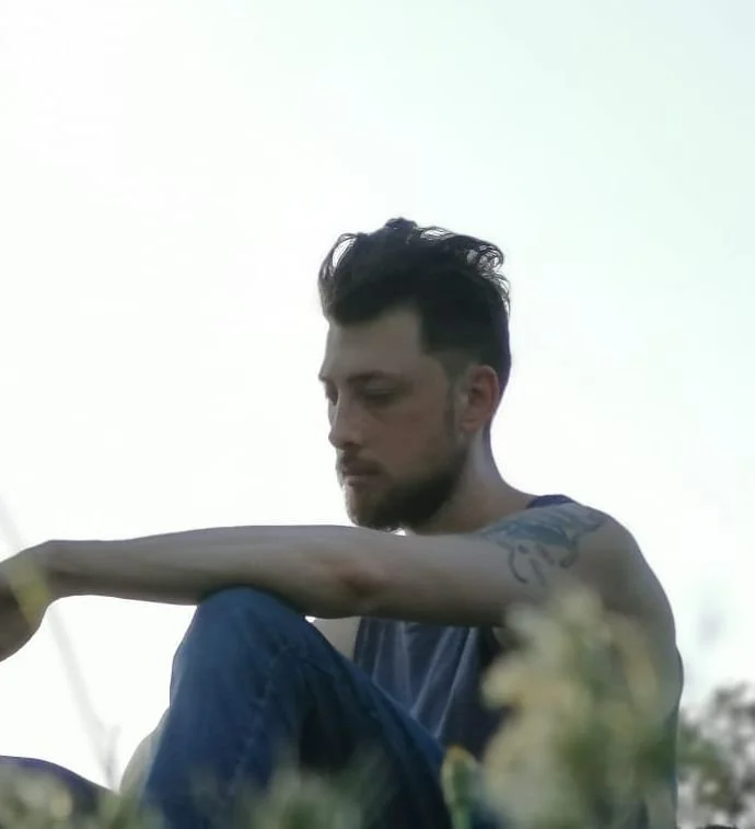 Jeune homme aux cheveux noirs et barbe, assis en extérieur, regardant vers le bas, dans un environnement naturel.
