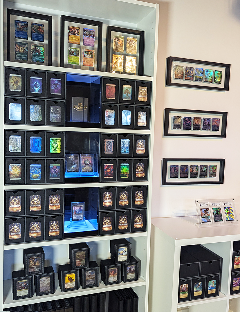 Espace d'exposition avec des cartes de jeux de société ou cartes à collectionner organisées dans des boîtiers noirs et affichées en cadre sur le mur.