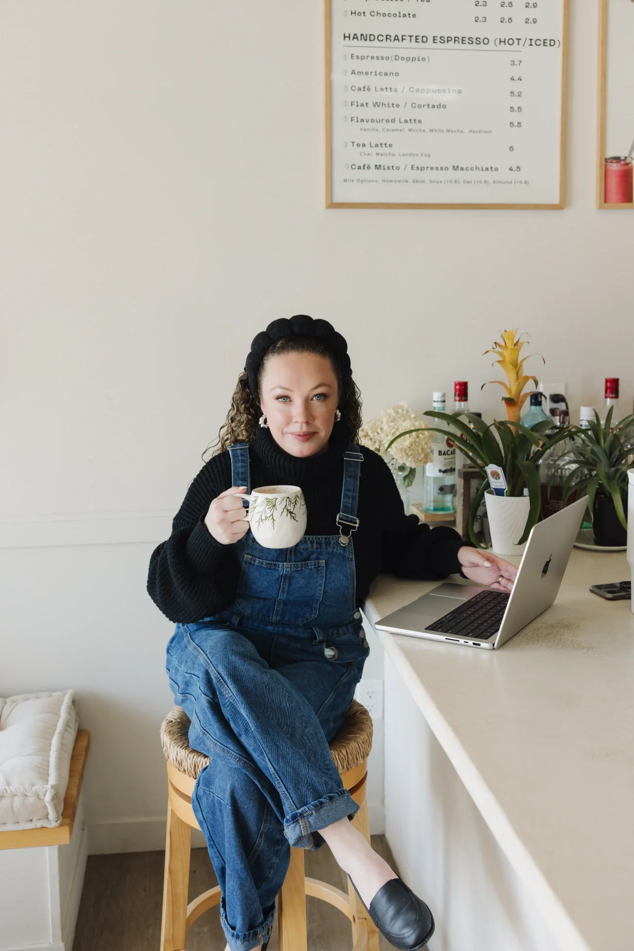 Cassie of Curl Media sitting on a wooden stool at a white counter, holding a cup of coffee in one hand and using her laptop with the other hand.