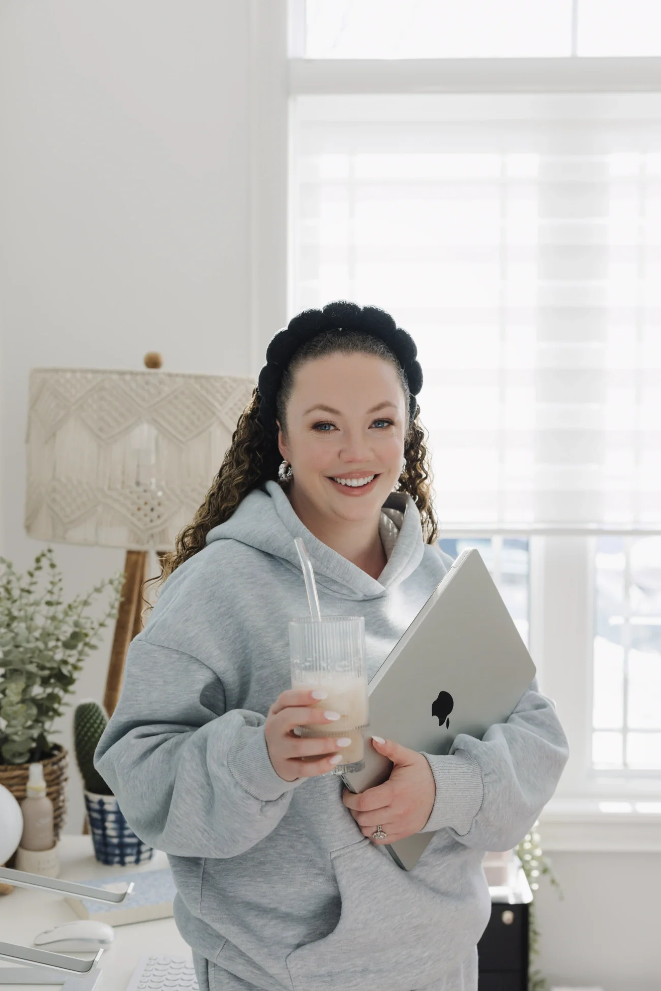 Cassie of Curl Media wearing a grey hoodie and black hairband smiling, holding a coffee in one hand and a laptop in the other.