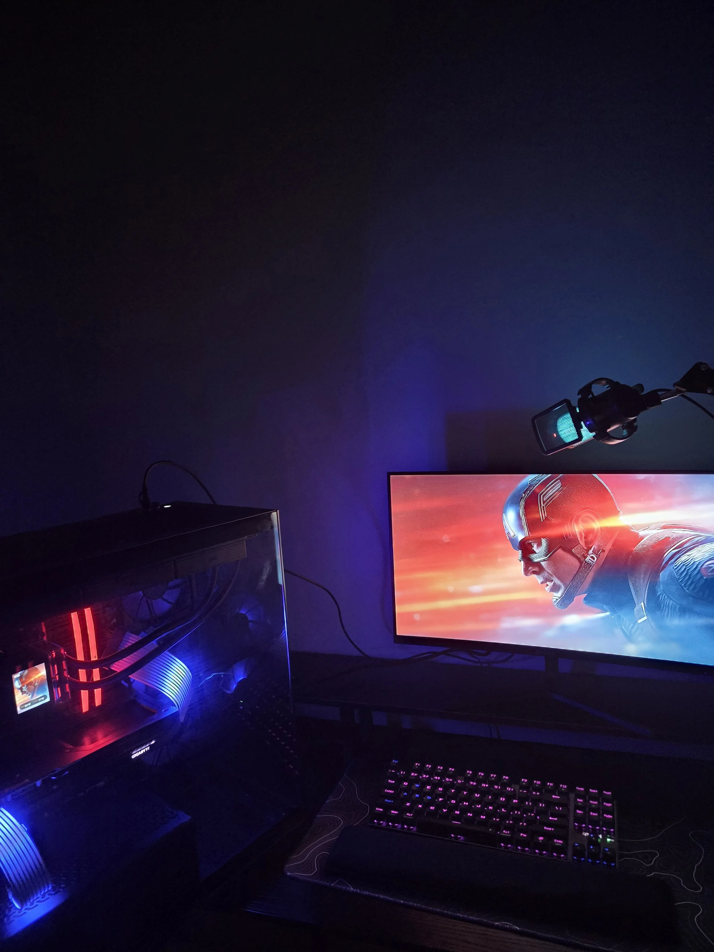 A gaming setup with a desktop computer with blue and red LED lights, a monitor displaying a superhero in a helmet with a red and blue background, a mechanical keyboard with RGB backlighting, and a gaming microphone mounted on a boom arm.
