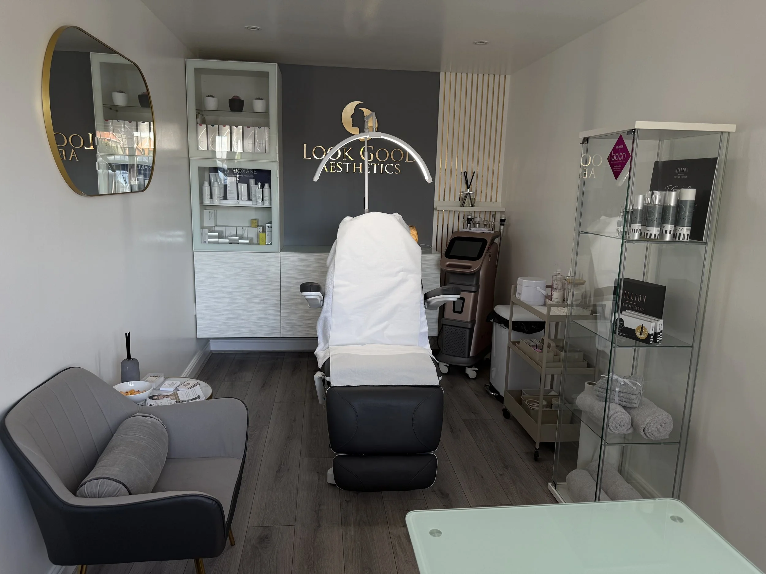 Interior of a beauty or aesthetic clinic with a treatment chair covered in white, a gray sofa with a pillow, glass display cases with skincare products, and various equipment.