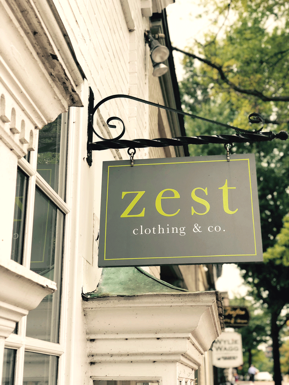 Storefront sign for 'zest clothing & co.' hanging on a black wrought iron bracket outside a building with white brick walls and large windows, with trees visible in the background.