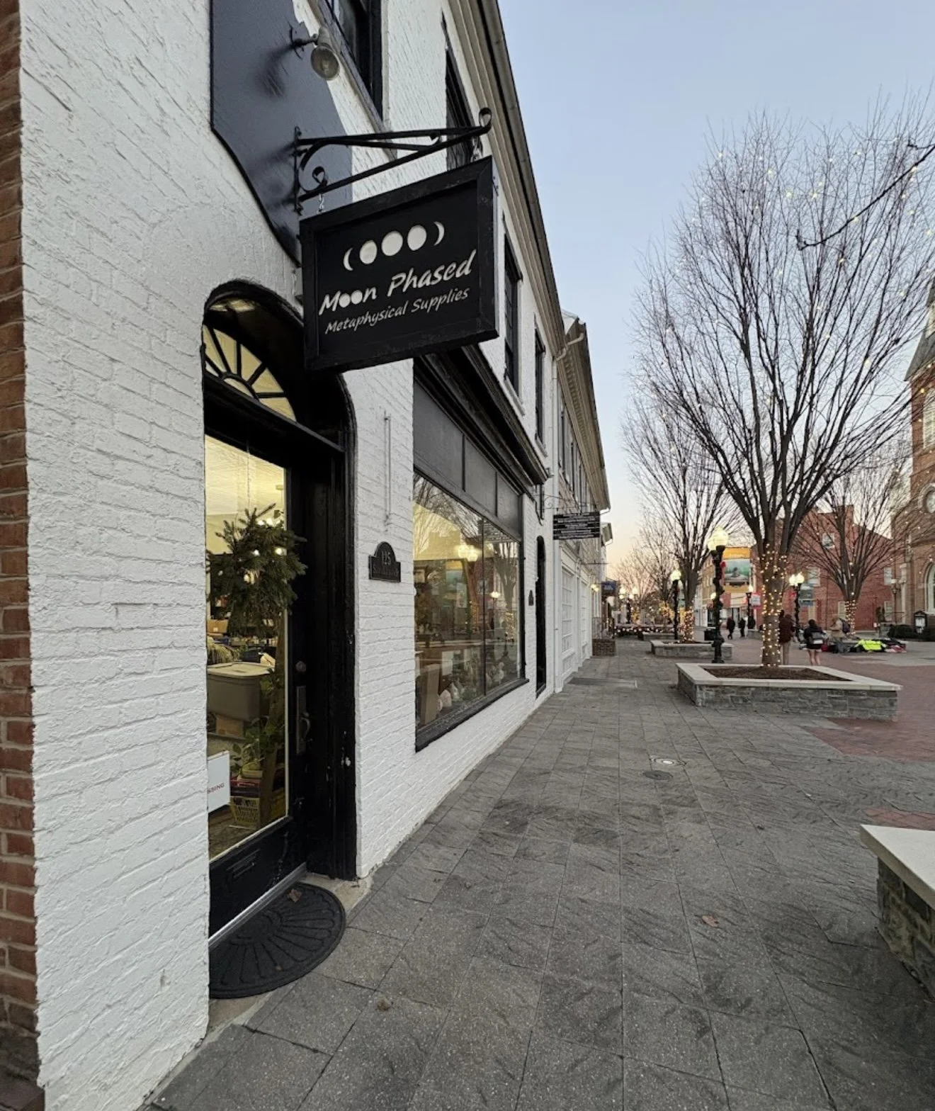 Street view of a shop called Moon Phased Metaphysical Supplies with a black sign, white painted brick wall, glass door, and large window showcasing shop interior, with leafless trees and streetlights along a quiet sidewalk during evening.