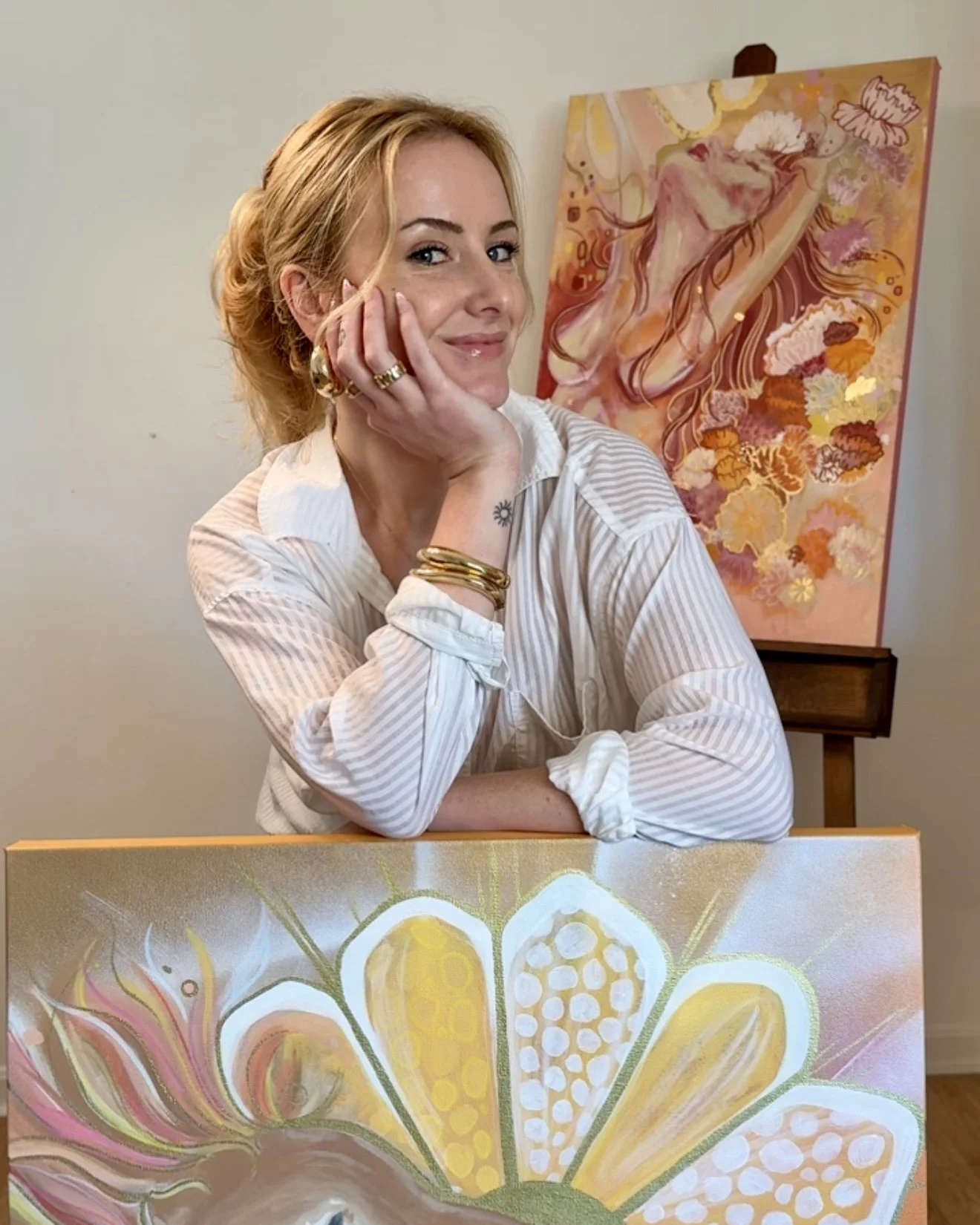 A woman with blonde hair smiling, resting her chin on her hand, wearing a white striped shirt and gold jewelry, with colorful abstract floral paintings in the background and foreground.