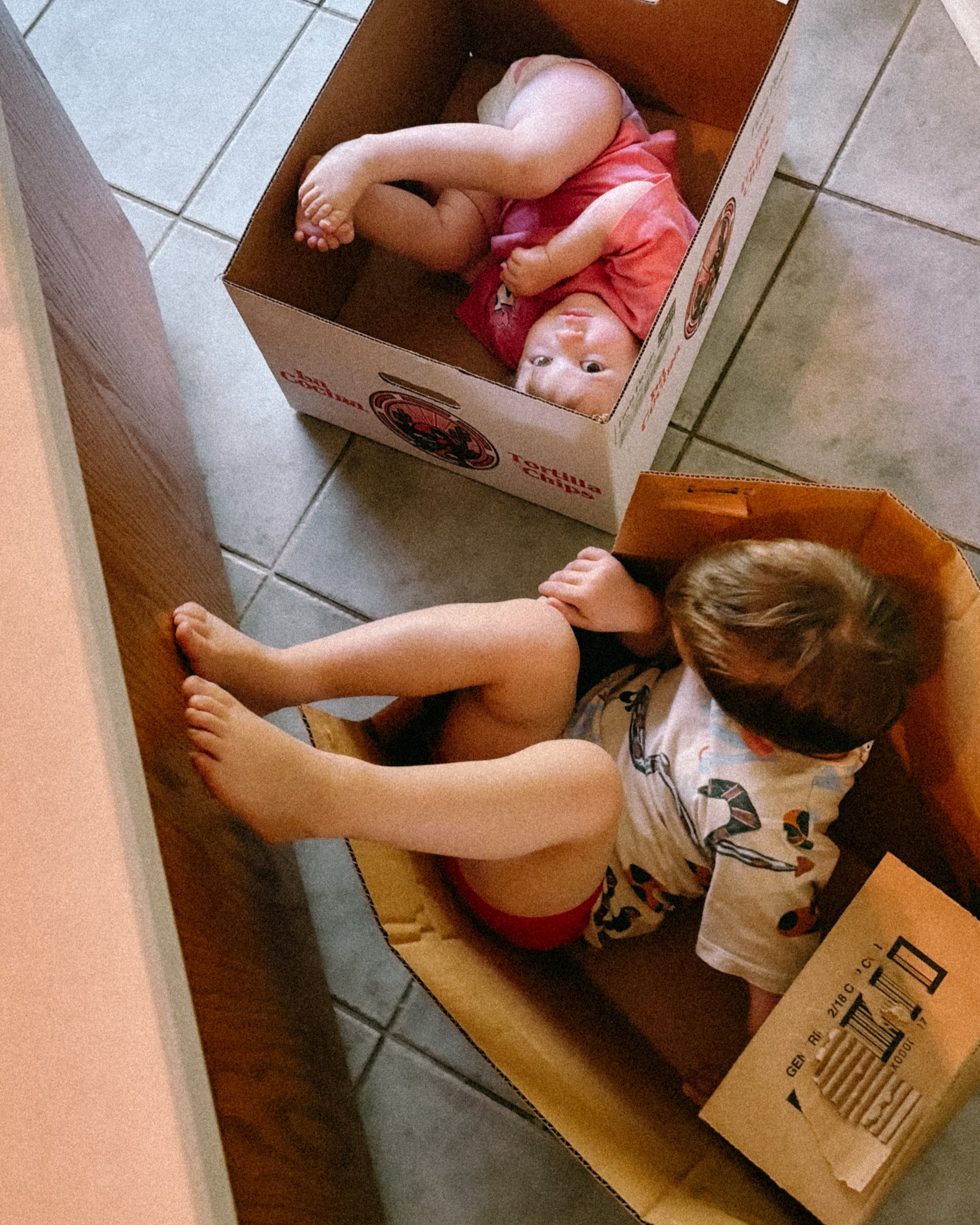 Two young children playing inside empty cardboard boxes on a tiled floor.