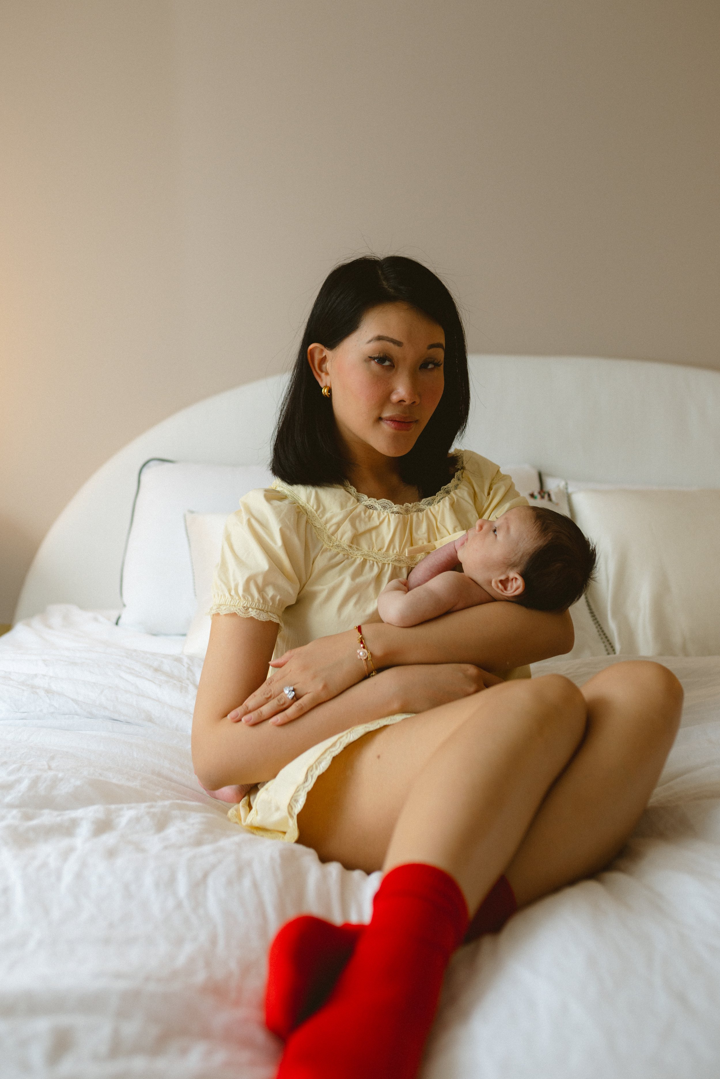 A woman with black hair, wearing a yellow dress, is sitting on a bed holding a baby in her arms. She looks at the baby with a gentle expression, and the baby is looking up at her. The bed has white sheets and a beige headboard, and the background is 