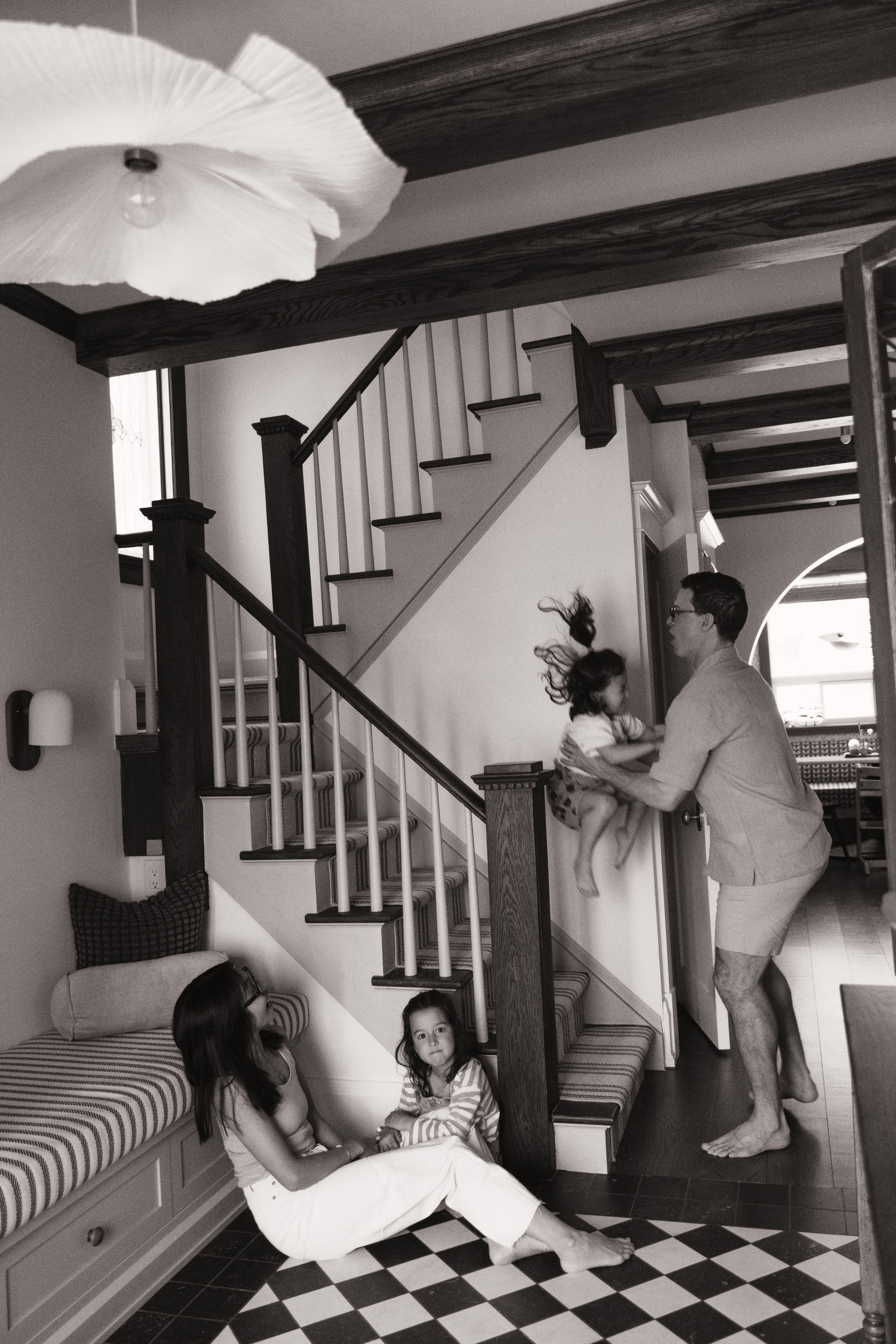 Black and white storytelling family photo of parents and children playing together near the stairs at home.