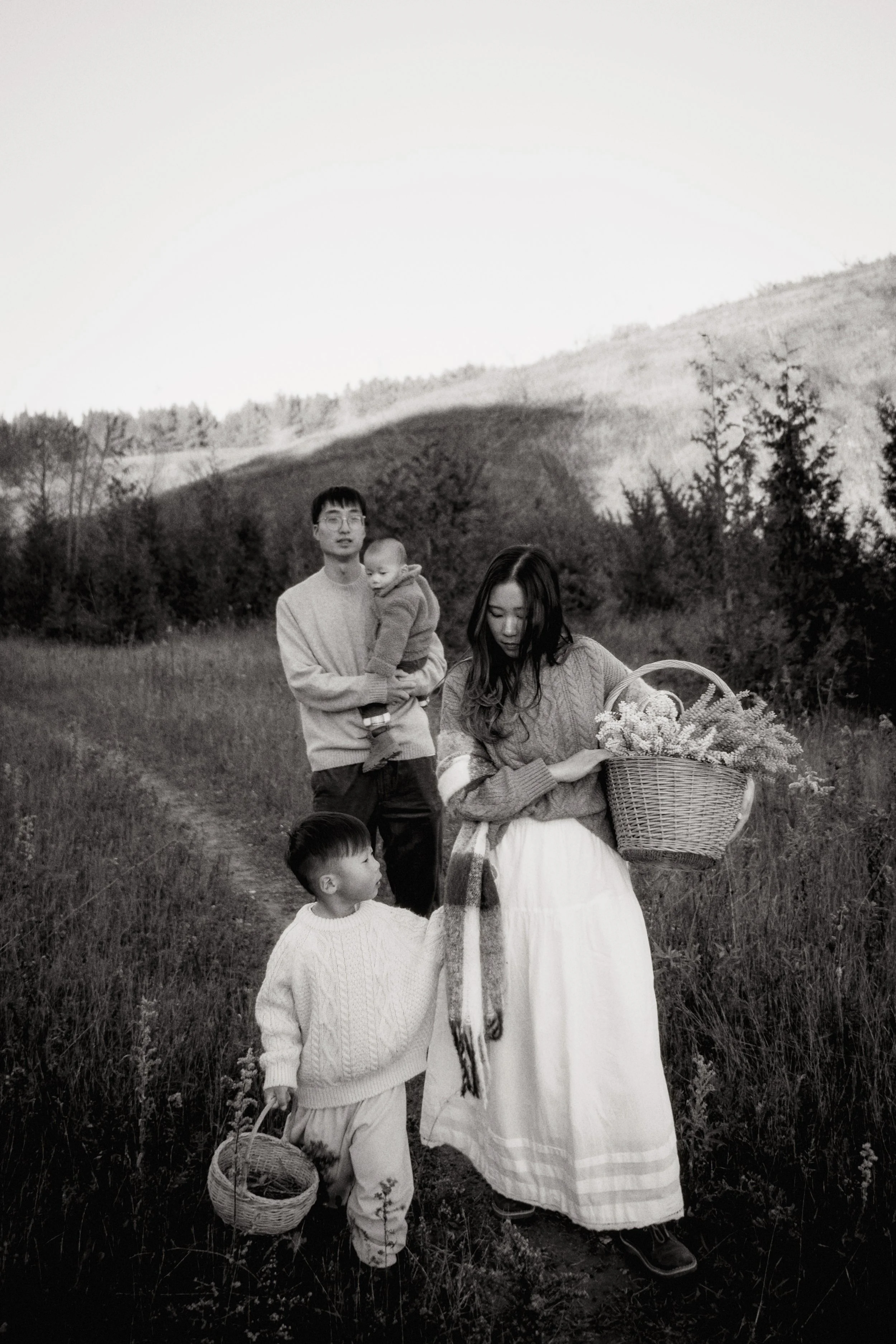 A family of four on a nature trail, with rolling hills and trees in the background. The woman is holding a basket of flowers, the man is holding a child, and a young boy is walking alongside her with a basket.