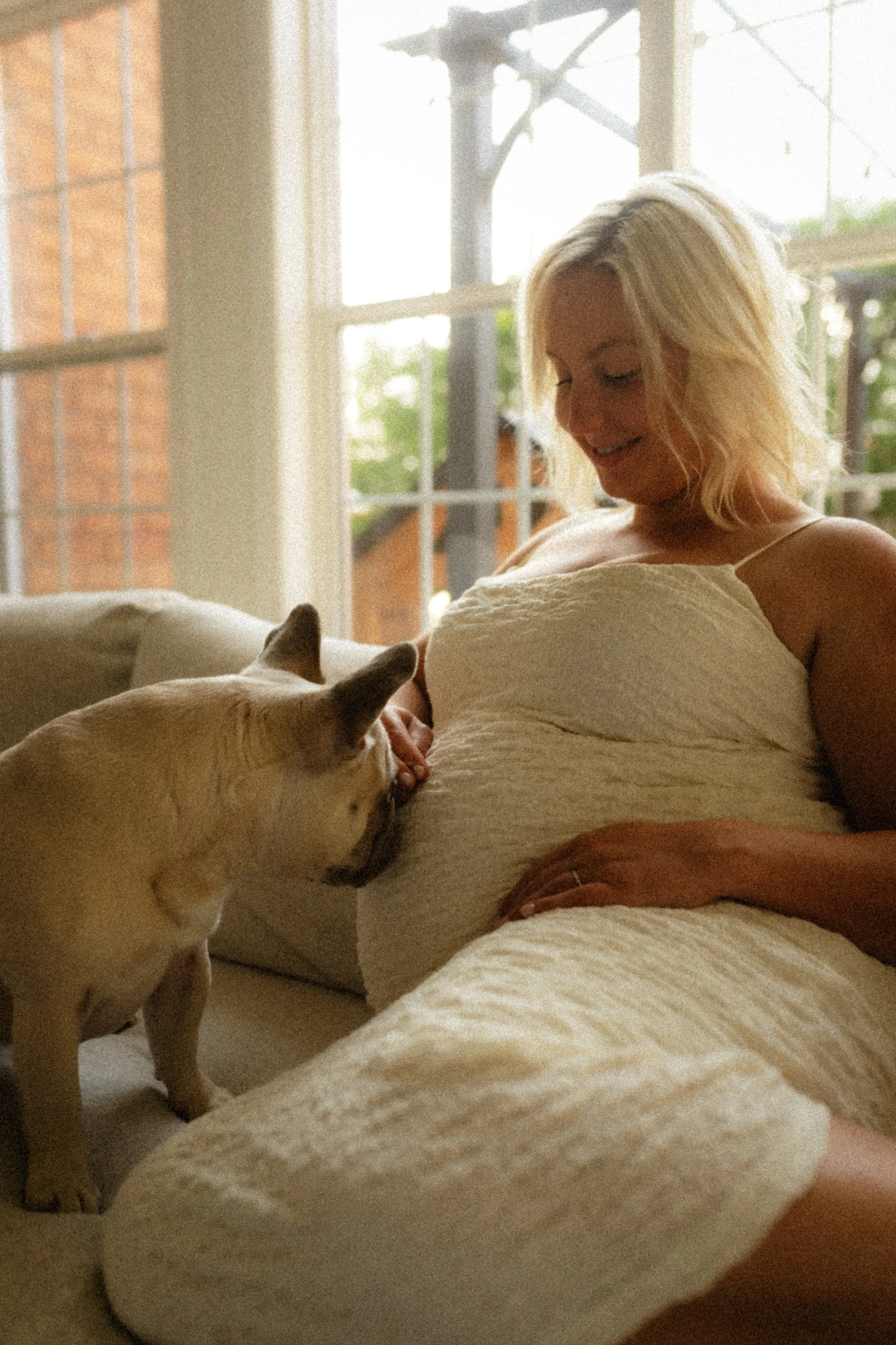 A woman sitting on a couch with a small dog sitting on a cushion, indoors near a large window with natural light.