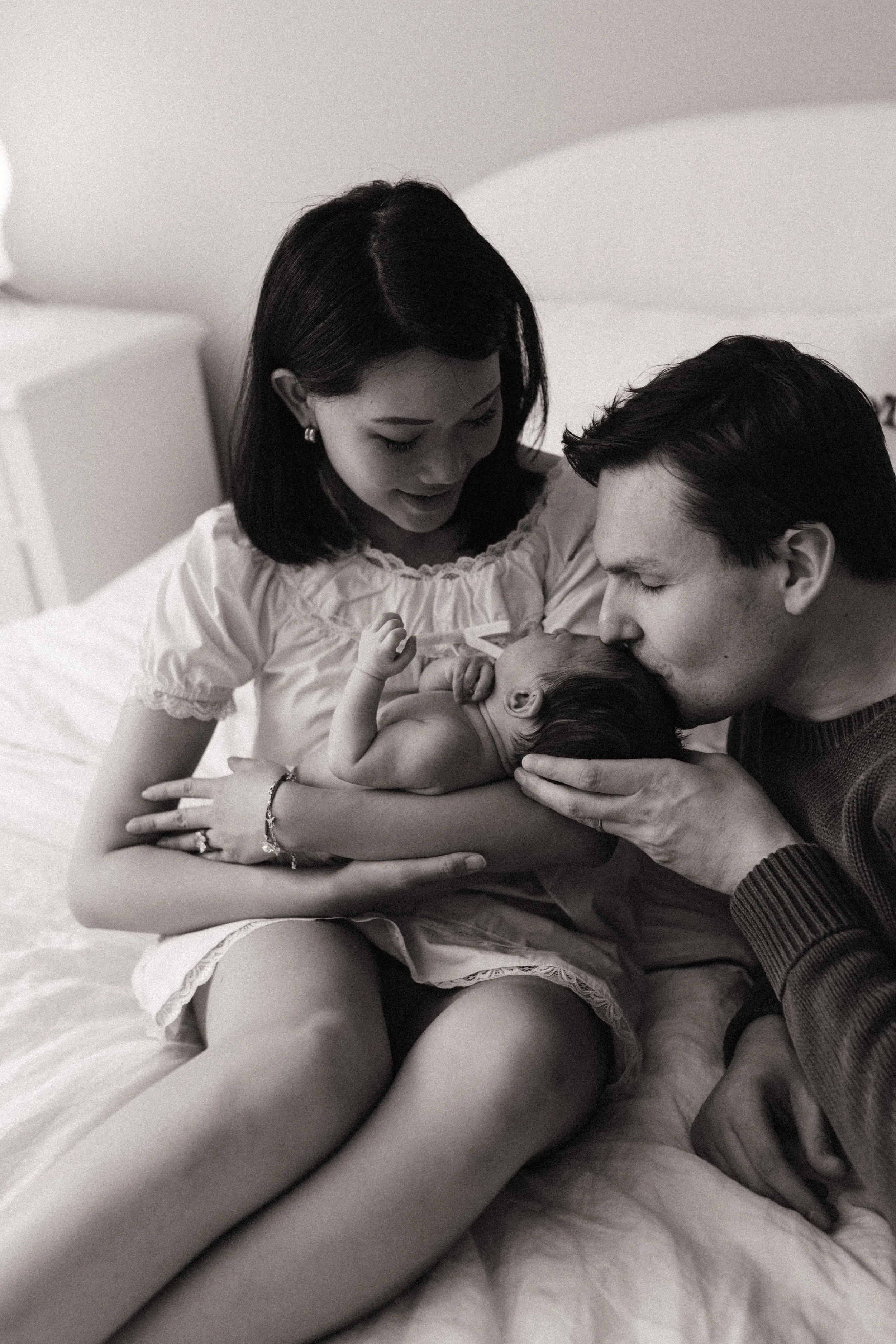 A black and white photo of a woman, a man, and a baby on a bed. The woman and man are looking at the newborn baby, who is lying on the woman's lap. The woman wears earrings and a bracelet, and the man has short hair. They are all engaged in a tender moment.