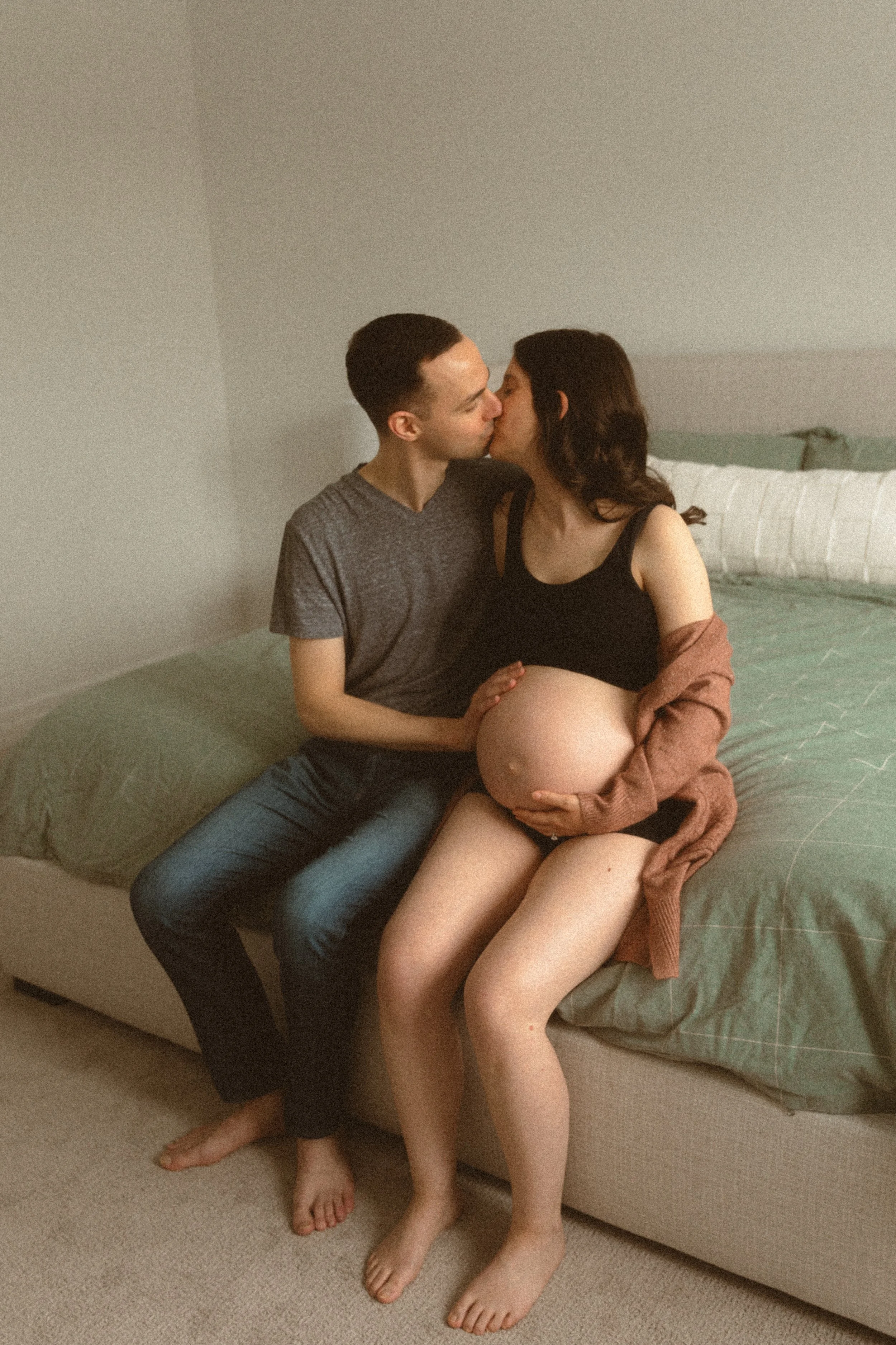 A man and a pregnant woman sitting on a bed sharing a kiss, with the woman holding her bare pregnant belly.