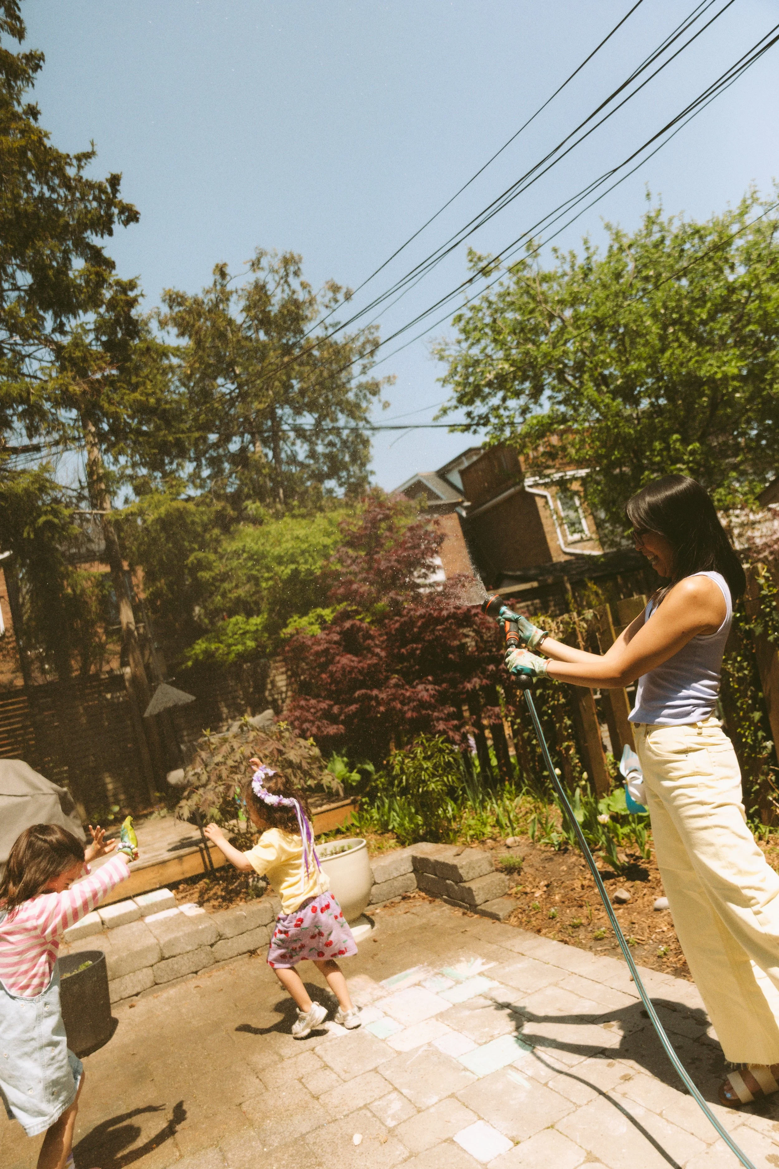 Candid backyard family photo of children playing with water while their mother holds the hose during an at-home family session.