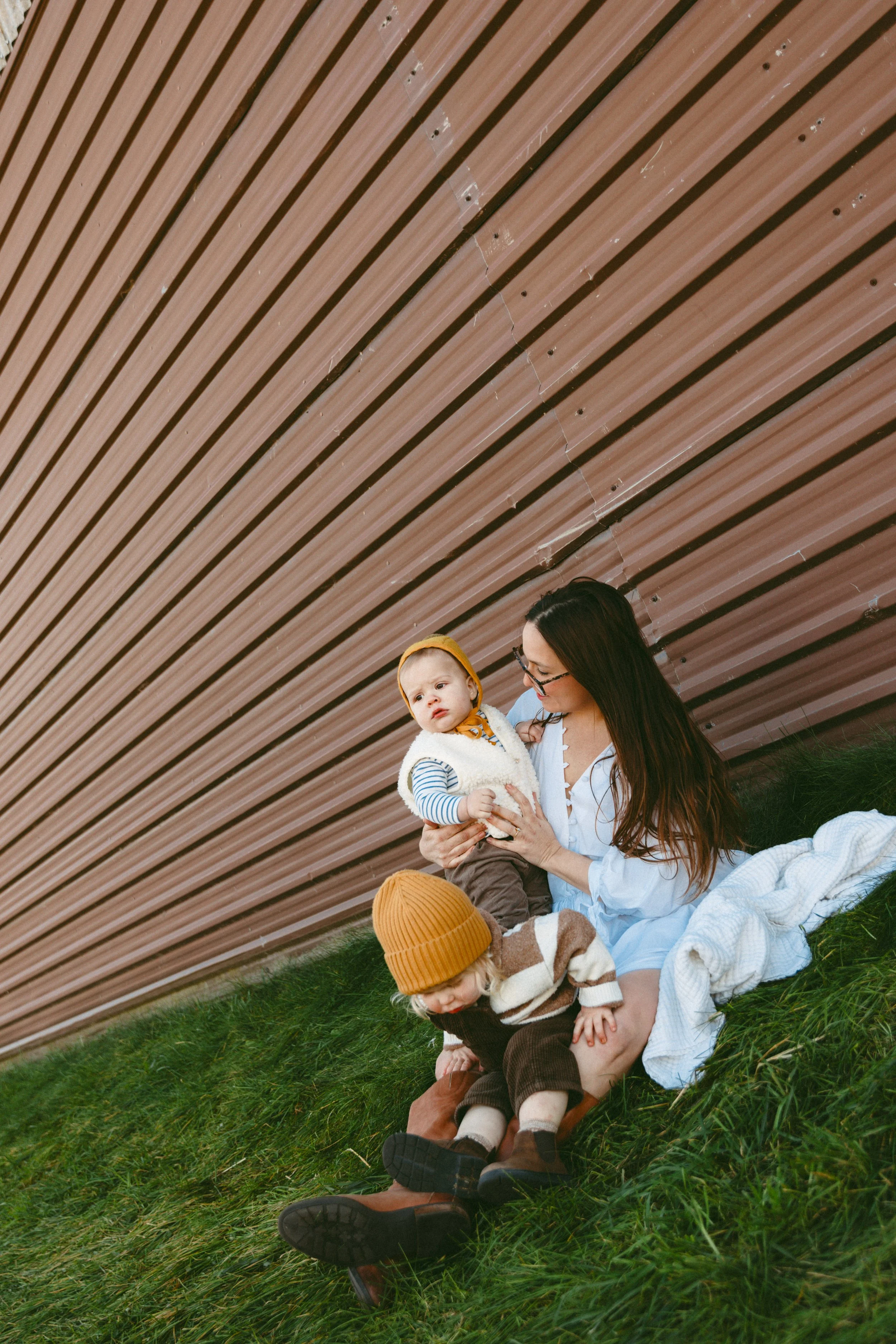 A woman with long brown hair and glasses holding a young child with a yellow headband, sitting on grass under a curved, brown wooden roof structure with two young children, one in a striped shirt and a yellow hat, and the other in a brown and white s