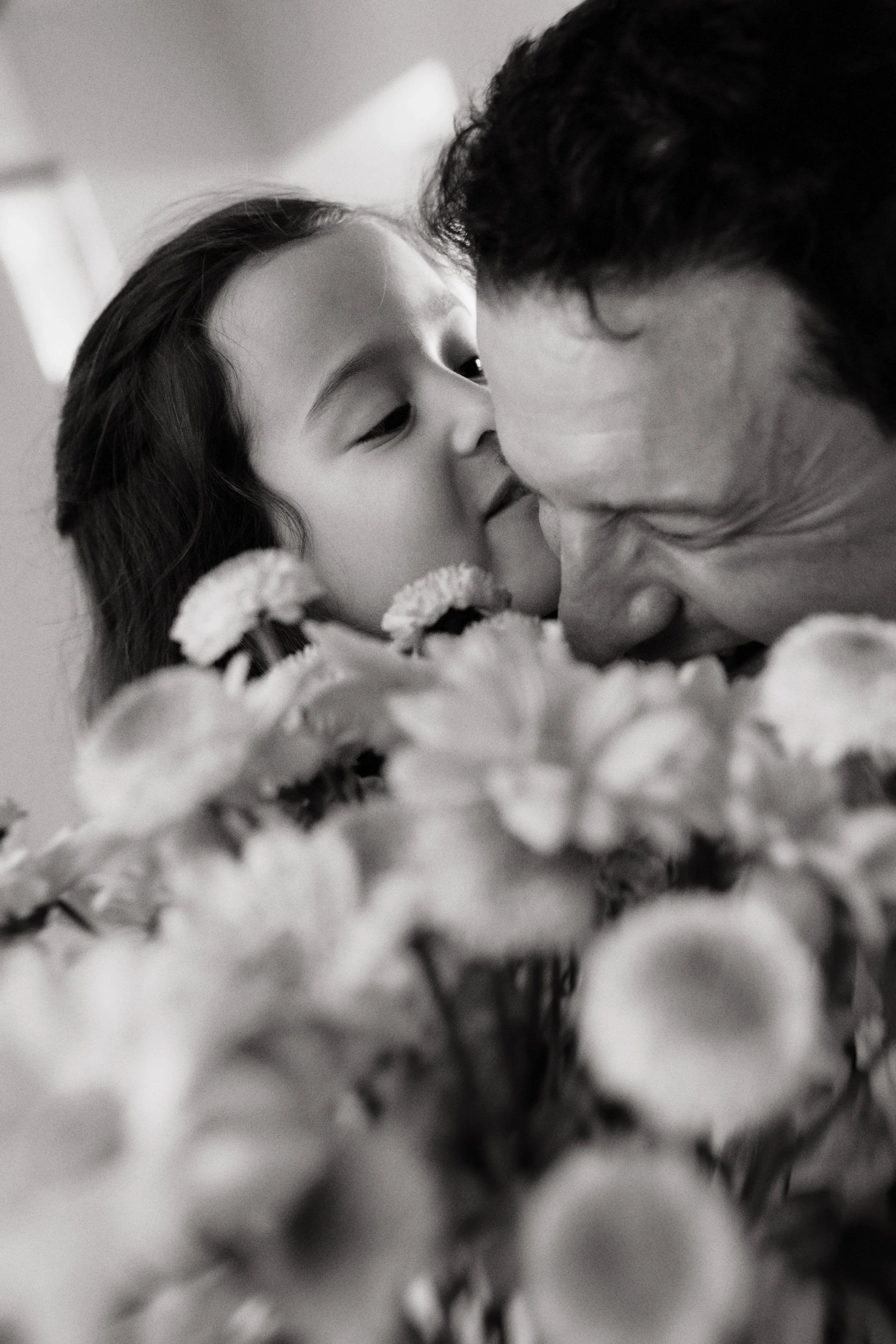 Black and white close-up of a father and daughter laughing together behind flowers during a candid family session at home.