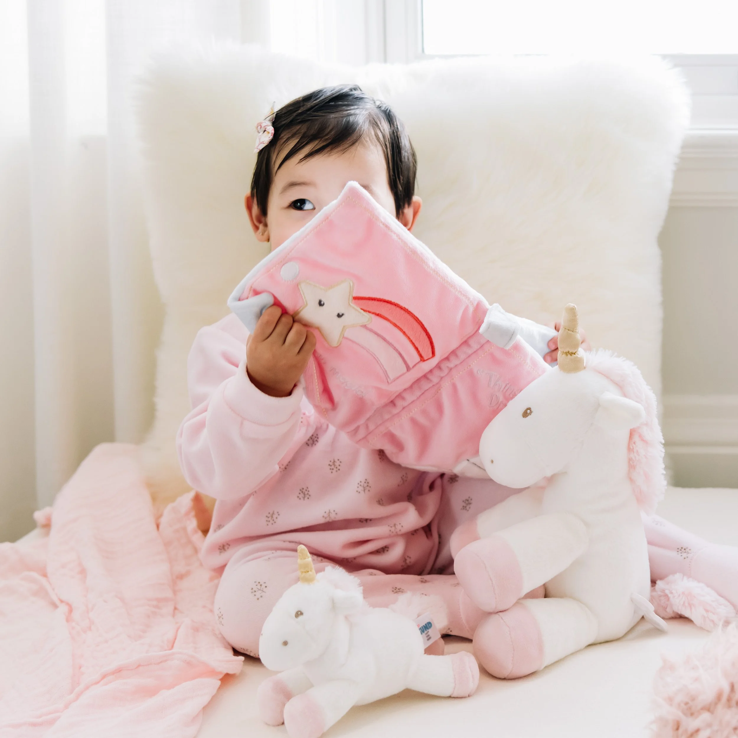 A young child with dark hair wearing a pink hair bow and holding a pink blanket with a star applique over a light pink blanket, sitting on a soft, pink fuzzy blanket near a window.