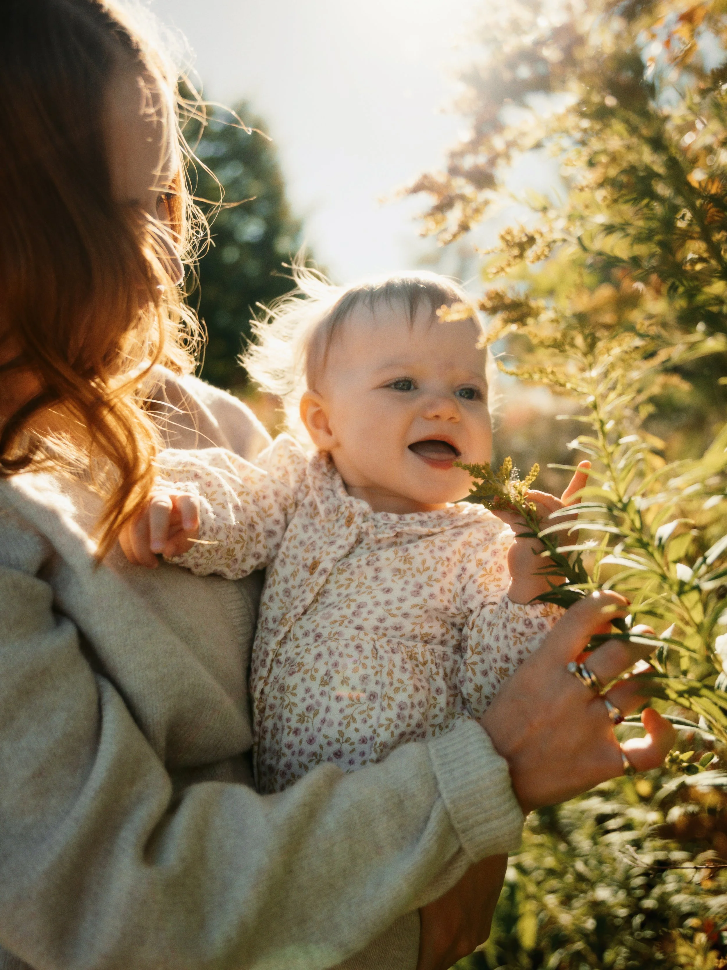 A woman holding a smiling baby girl outdoors in a garden with sunlight shining through trees, as the baby reaches for a plant.
