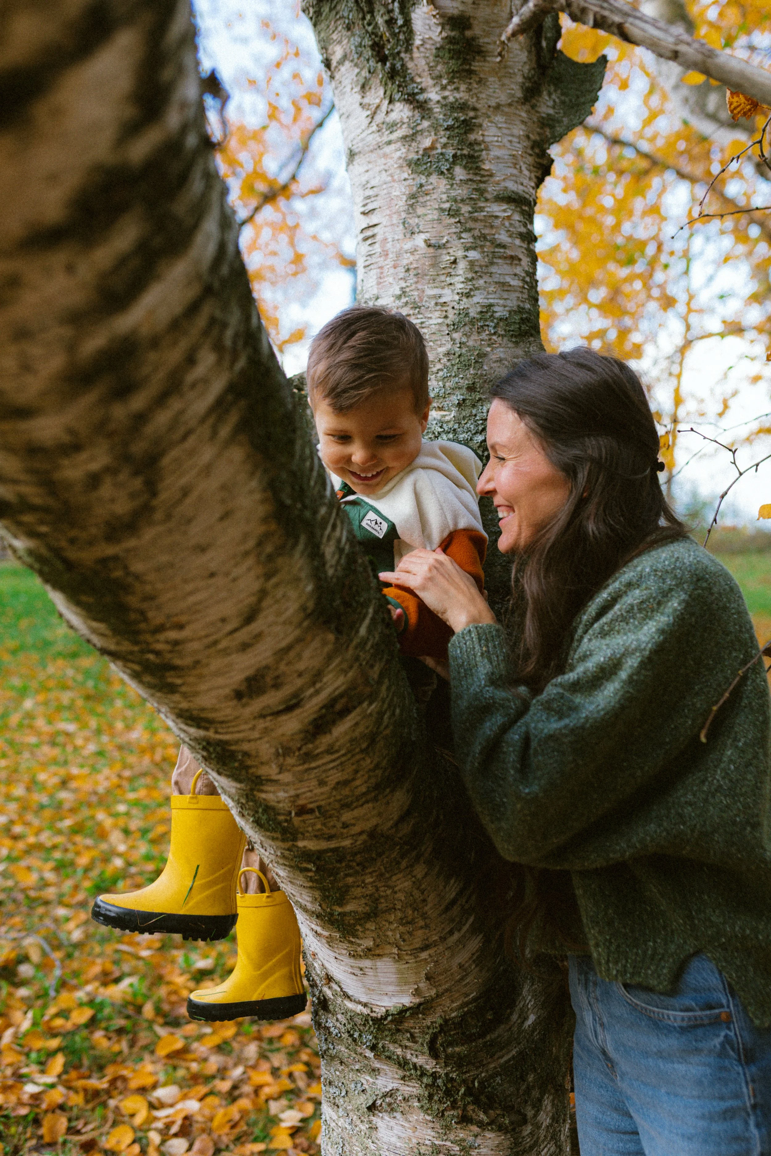 A woman and a young boy smiling and climbing a large tree with yellow boots, surrounded by autumn leaves.