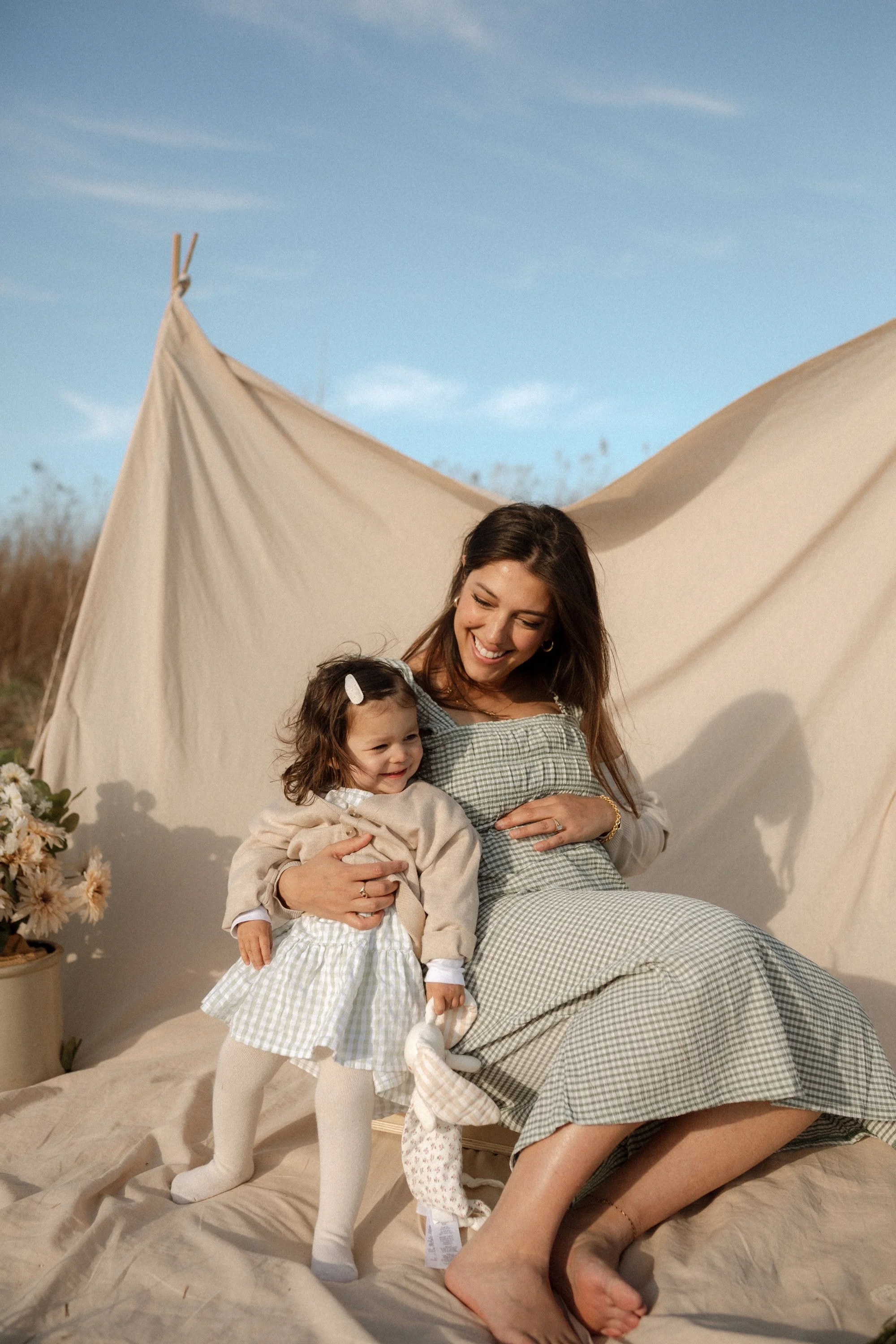 A woman and a young girl, smiling and hugging, sitting outdoors on a beige blanket in front of a beige tent under a blue sky.