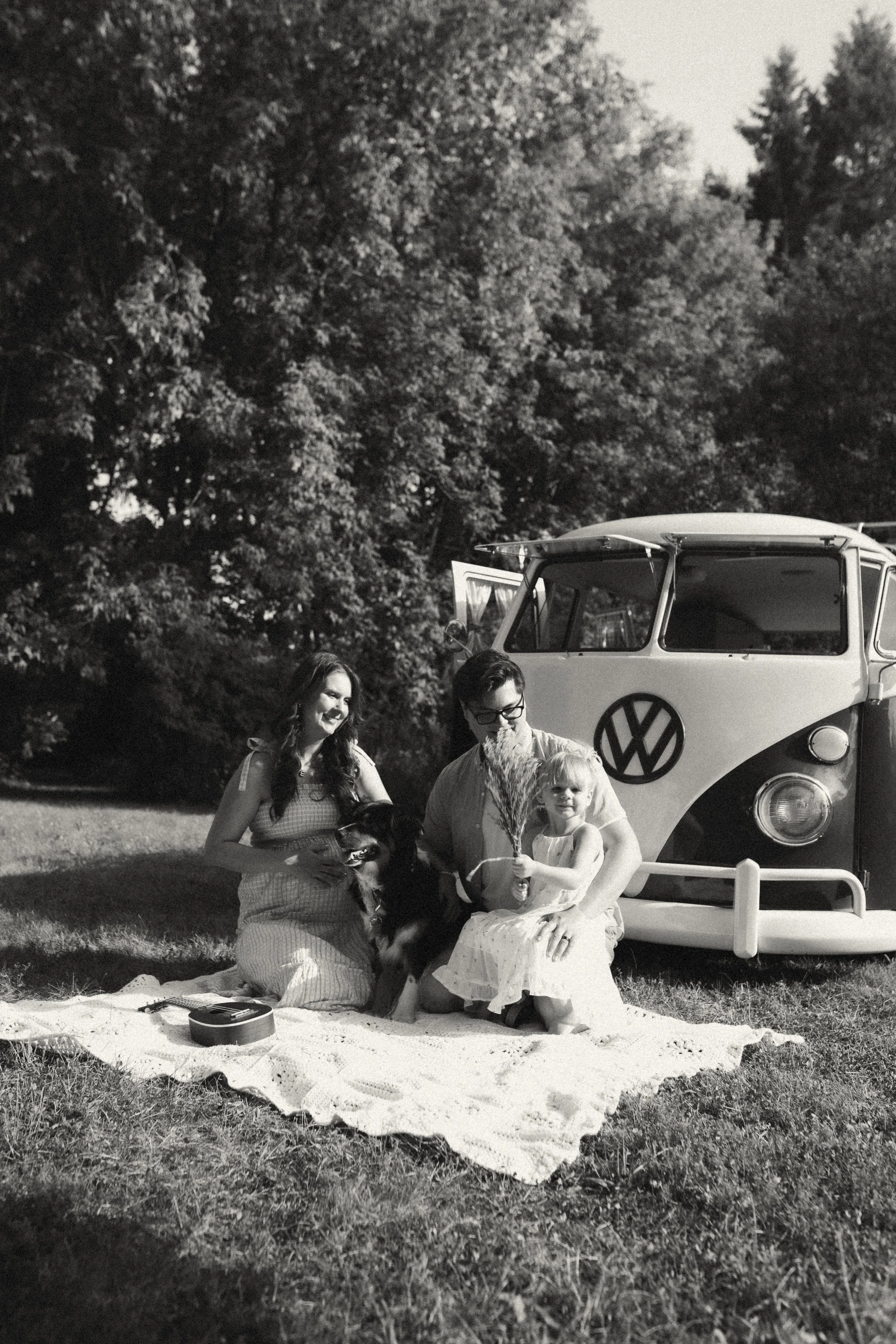 A black and white photo of three people and a dog sitting on a blanket outside near a vintage Volkswagen van, with trees in the background.