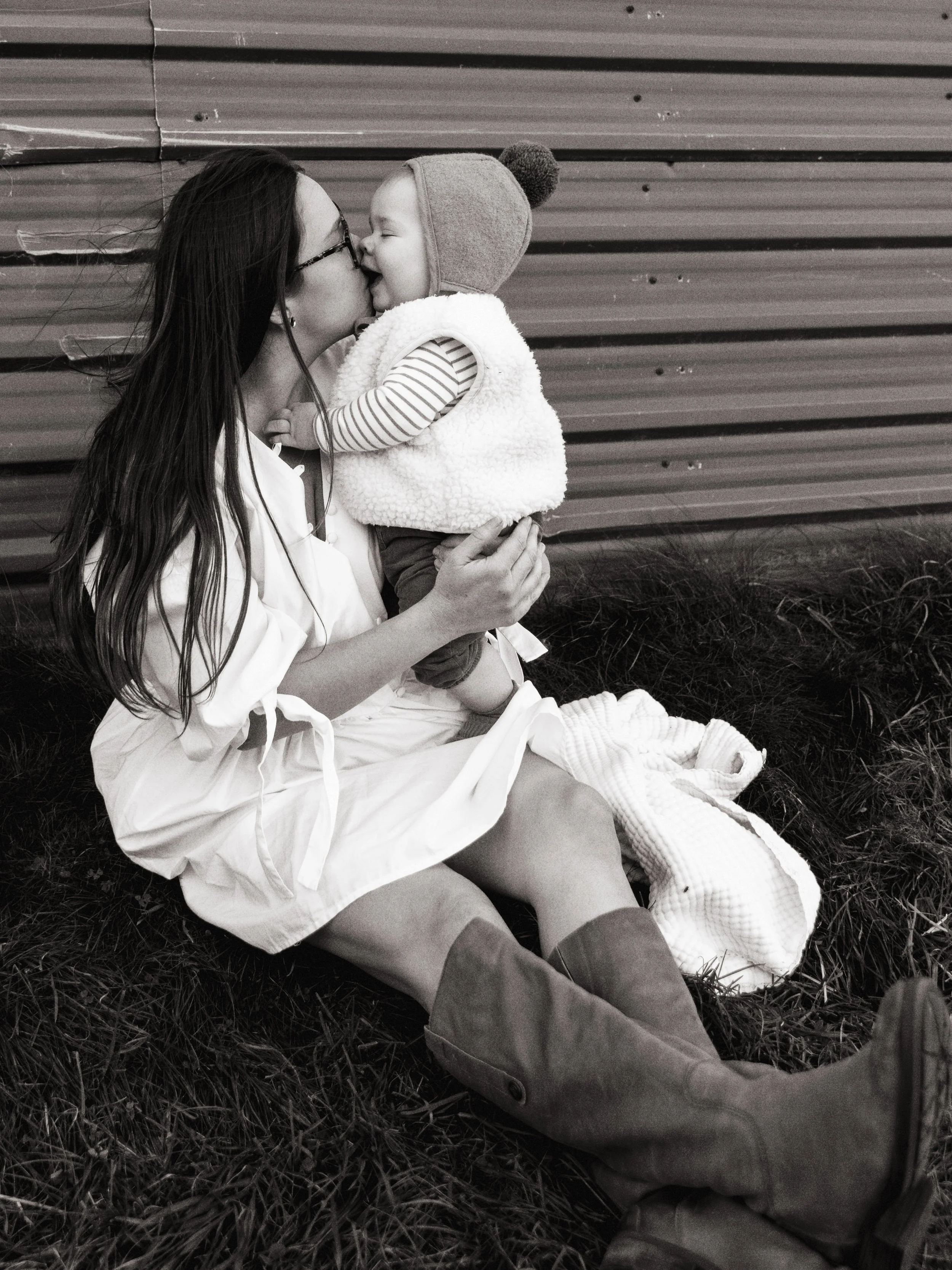 A woman with long hair and glasses holding a young child, both smiling and touching noses. They are sitting on grass near a corrugated metal background.