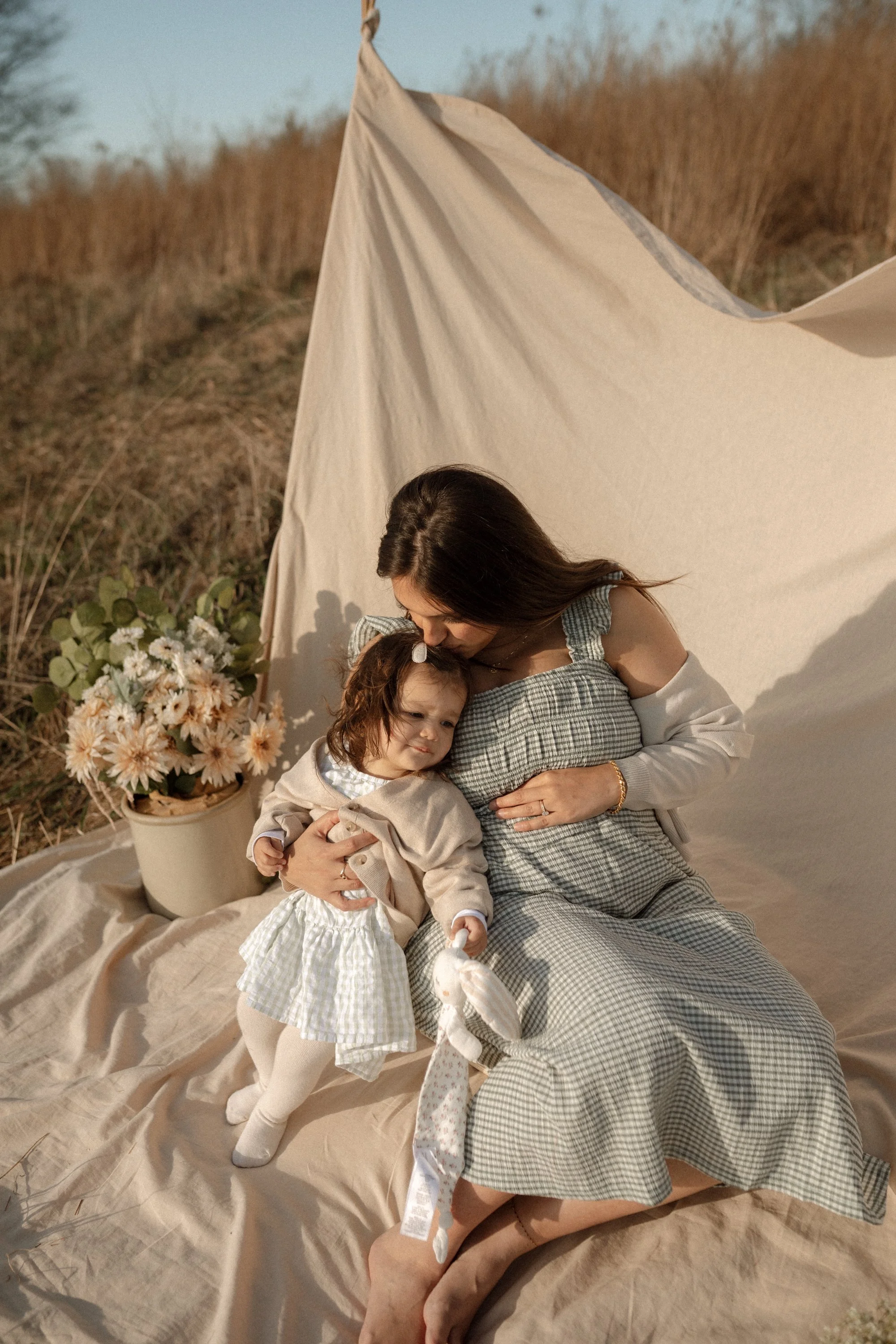 A woman and a young girl sitting outdoors on a beige blanket next to a teepee tent and a potted flower arrangement, sharing a tender moment.