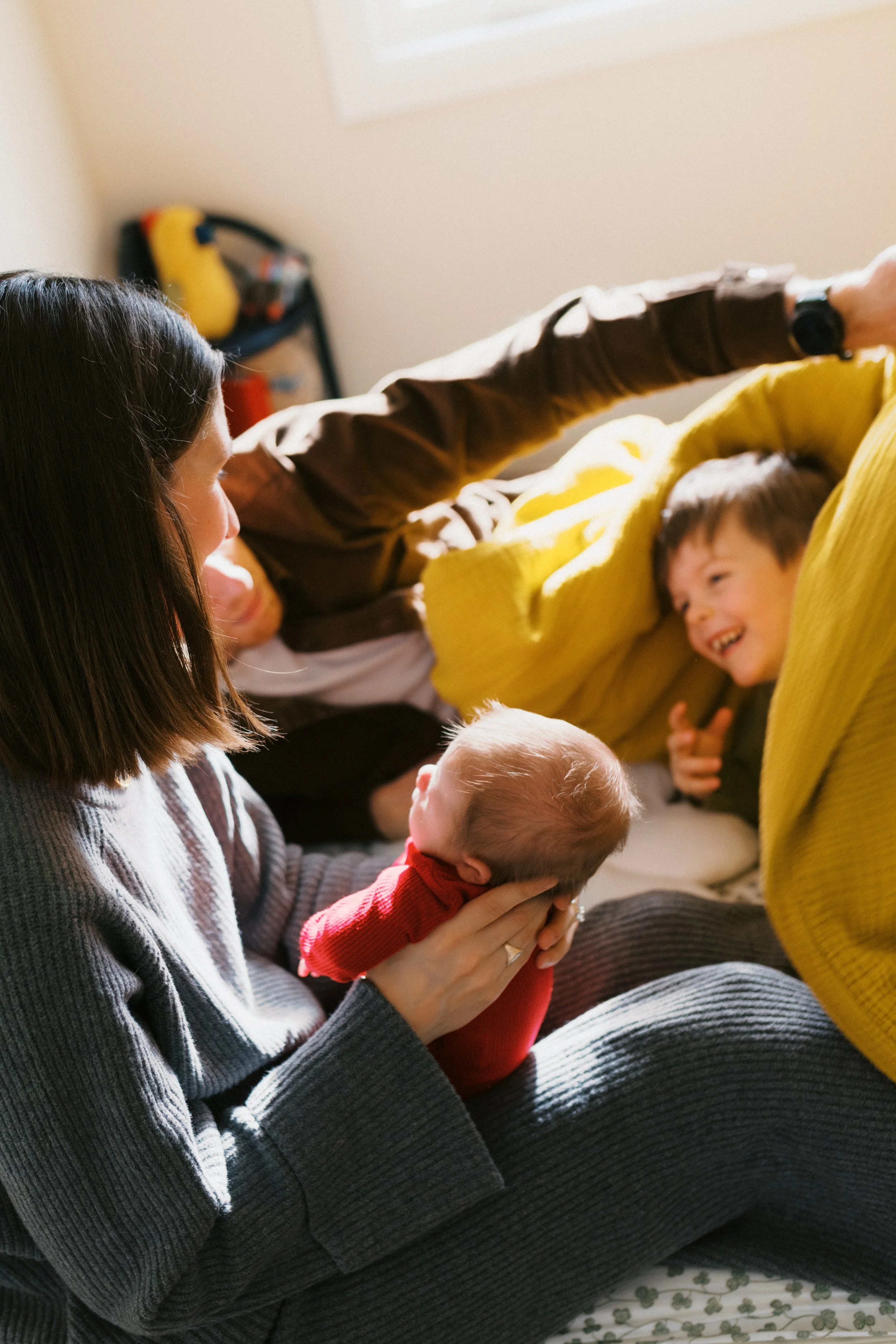A woman sitting on the floor holding a baby in her arms with a boy playing with her in a yellow sweater nearby. Sunlight coming through a window illuminates the scene.