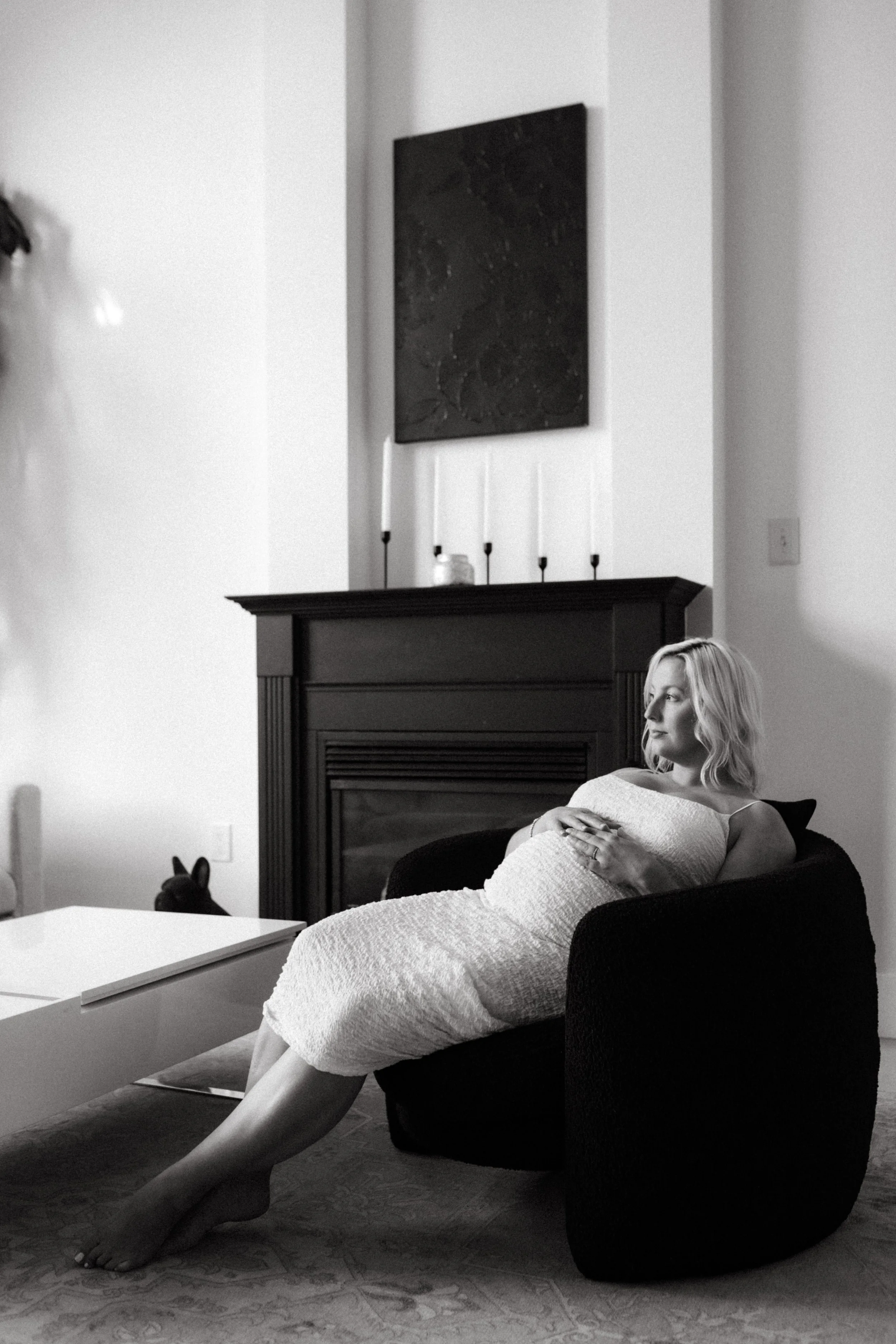 A woman sitting on a black armchair in a living room, holding a blanket or towel, with a decorative fireplace and candles on the mantel behind her, and a remote control in her hand.