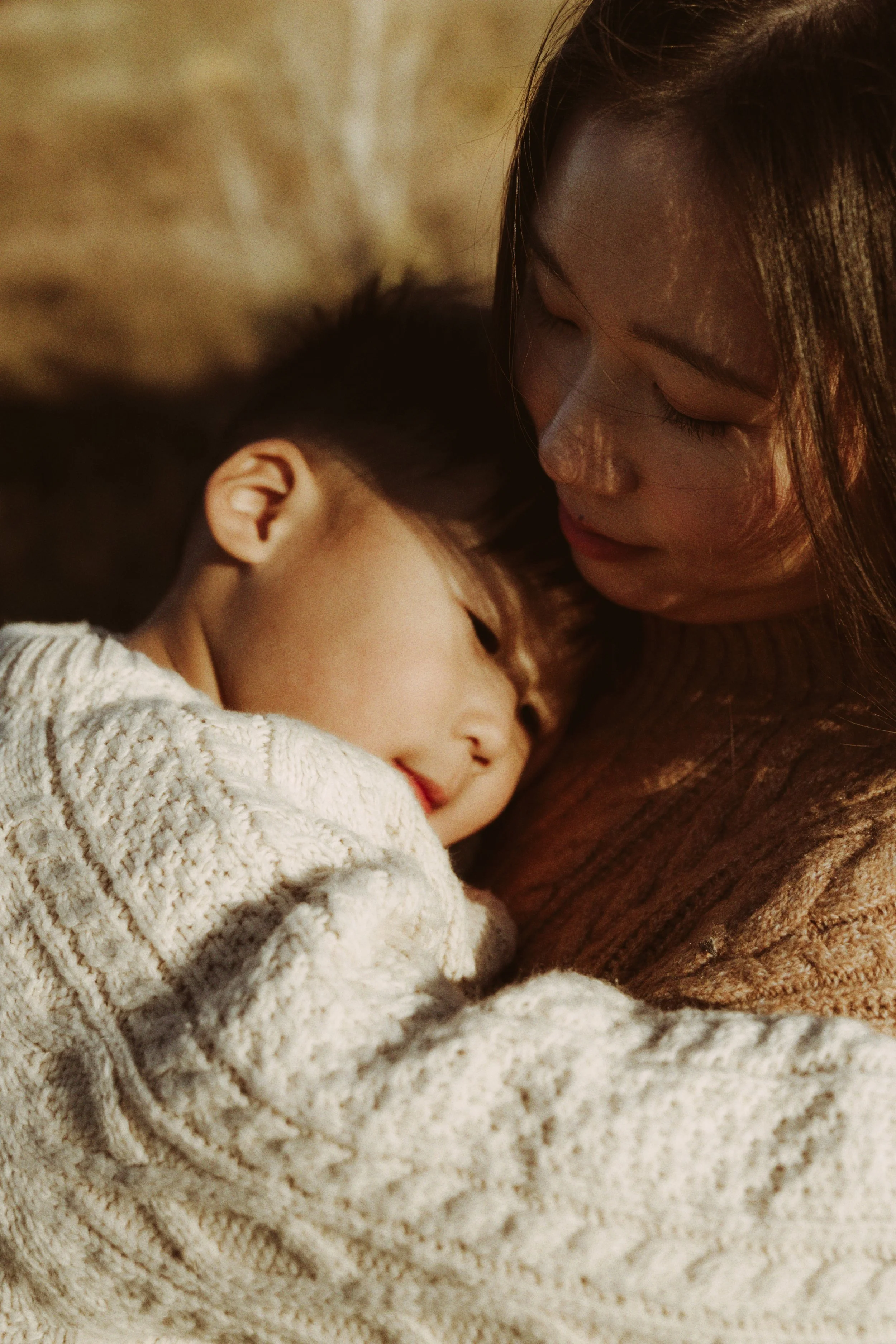 A woman gently holding and comforting a young girl resting her head on her shoulder, both with closed eyes, in a warm, cozy setting.