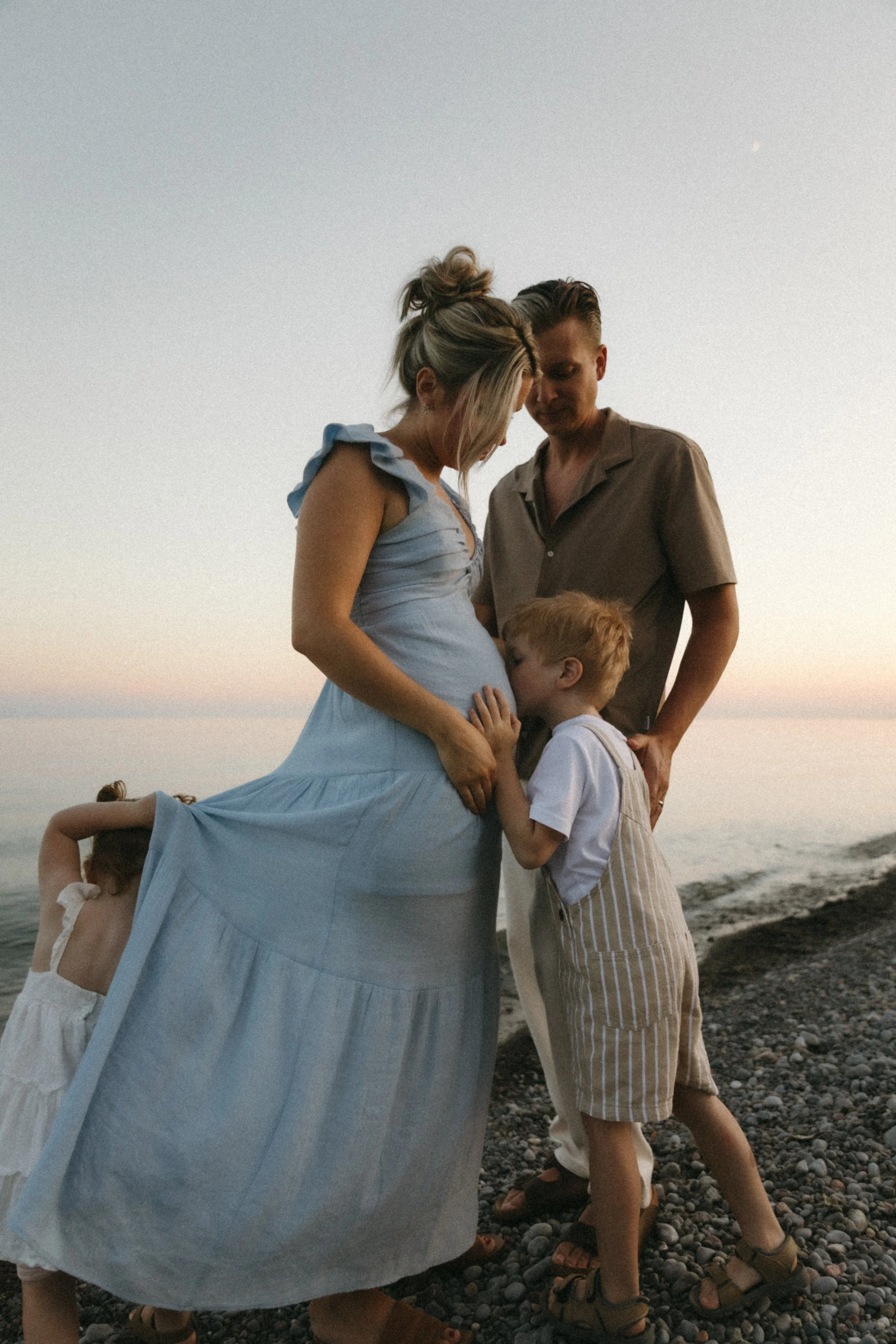 A pregnant woman with her family on a pebble beach at sunset, touching her belly as she stands with a man and two children, one of whom is kissing her belly.