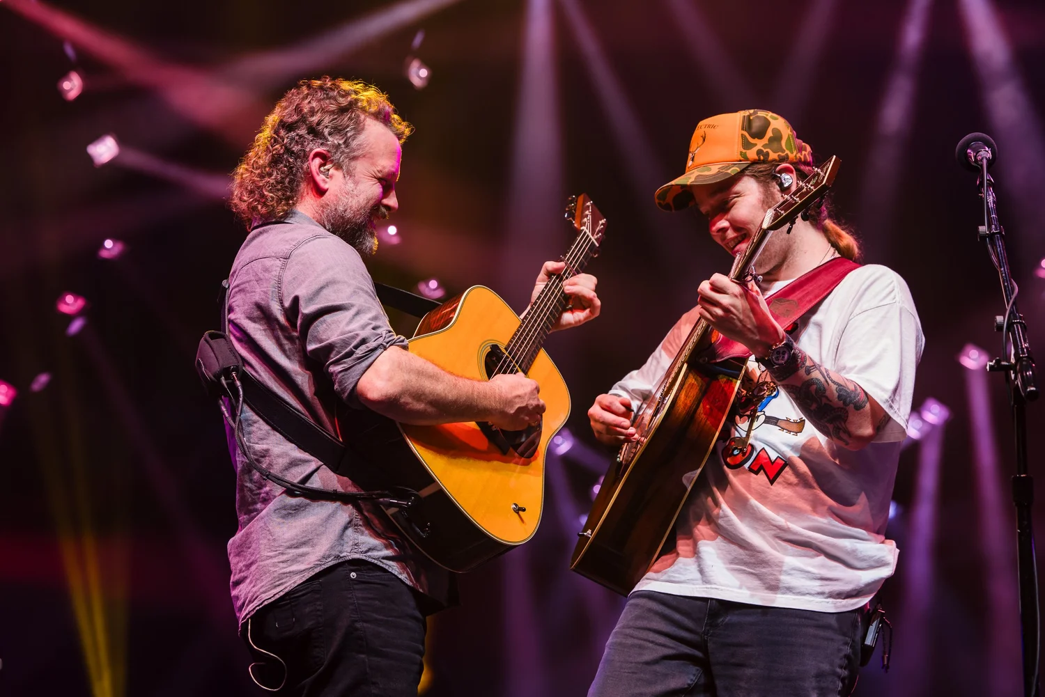 Jon Stickley playing acoustic guitar on stage alongside a fellow guitarist, both leaning in mid-performance under stage lighting