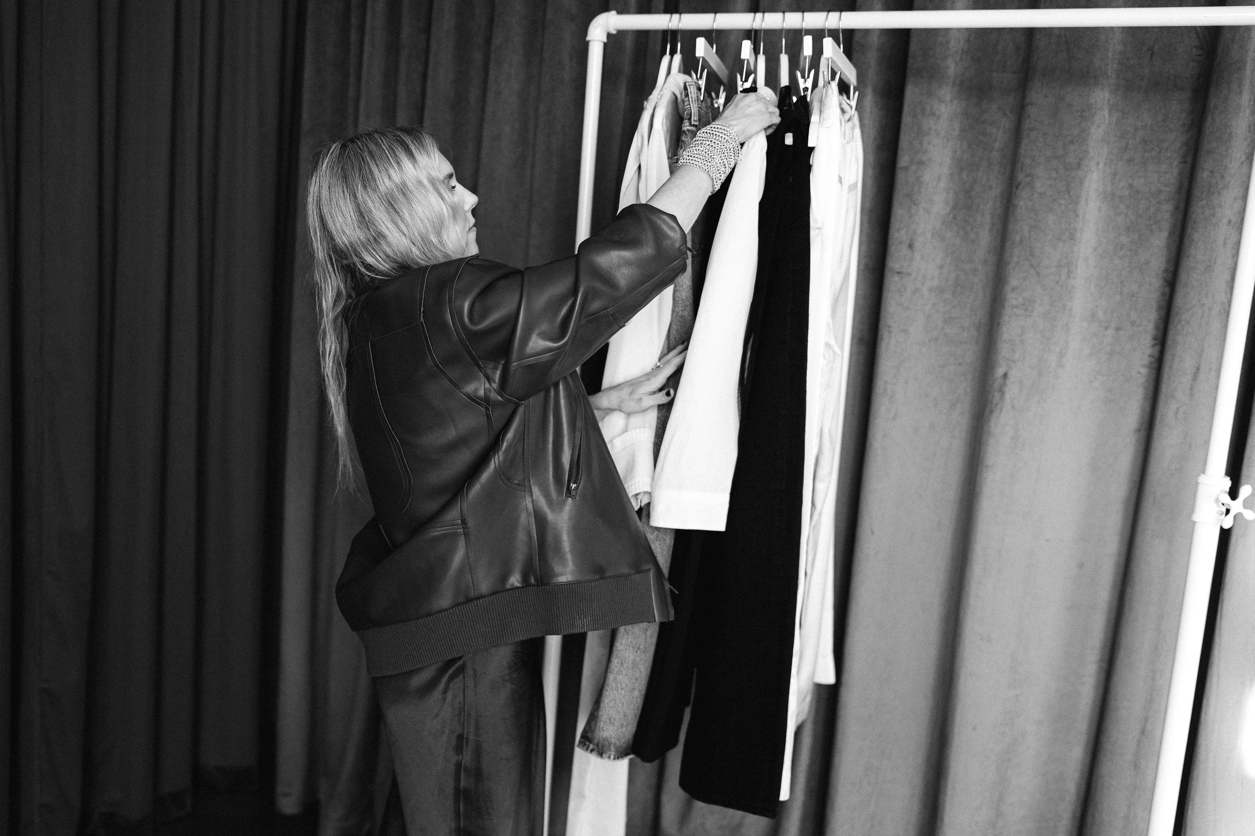 A woman browsing clothes on a rack, wearing a leather jacket and a wrist covering, in a fitting room or backstage environment.