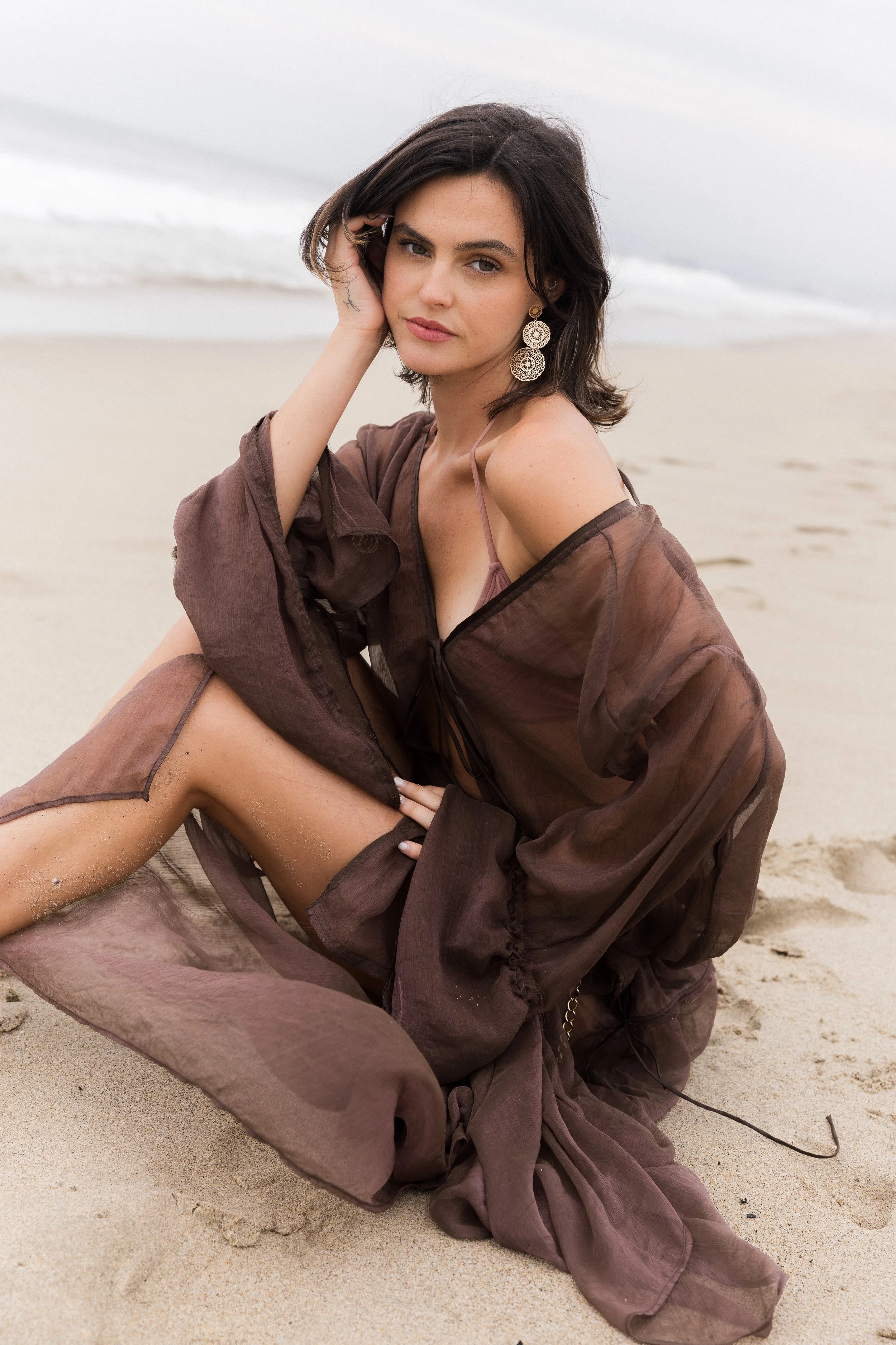 A woman with short dark hair and large earrings sitting on the sandy beach, wearing a sheer brown cover-up over a swimsuit, with the ocean and cloudy sky in the background.