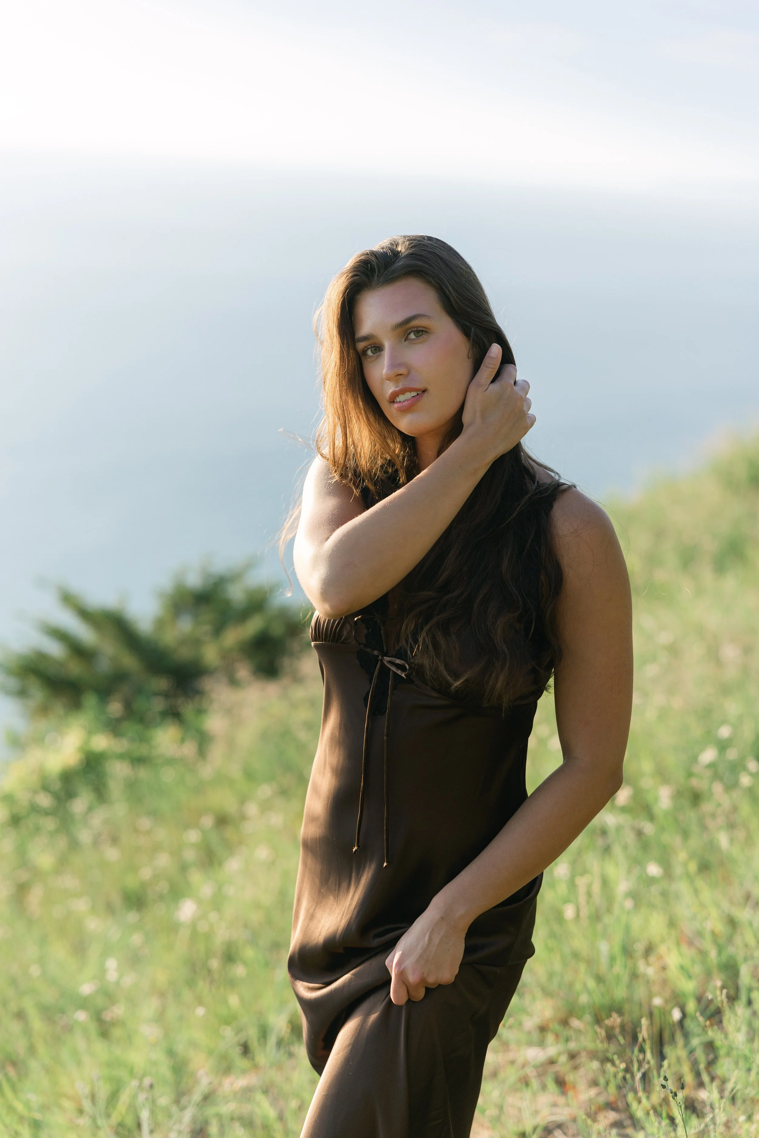 A woman with long brown hair standing outdoors in a green field with a blurred background of trees and sky, touching her hair with her right hand.