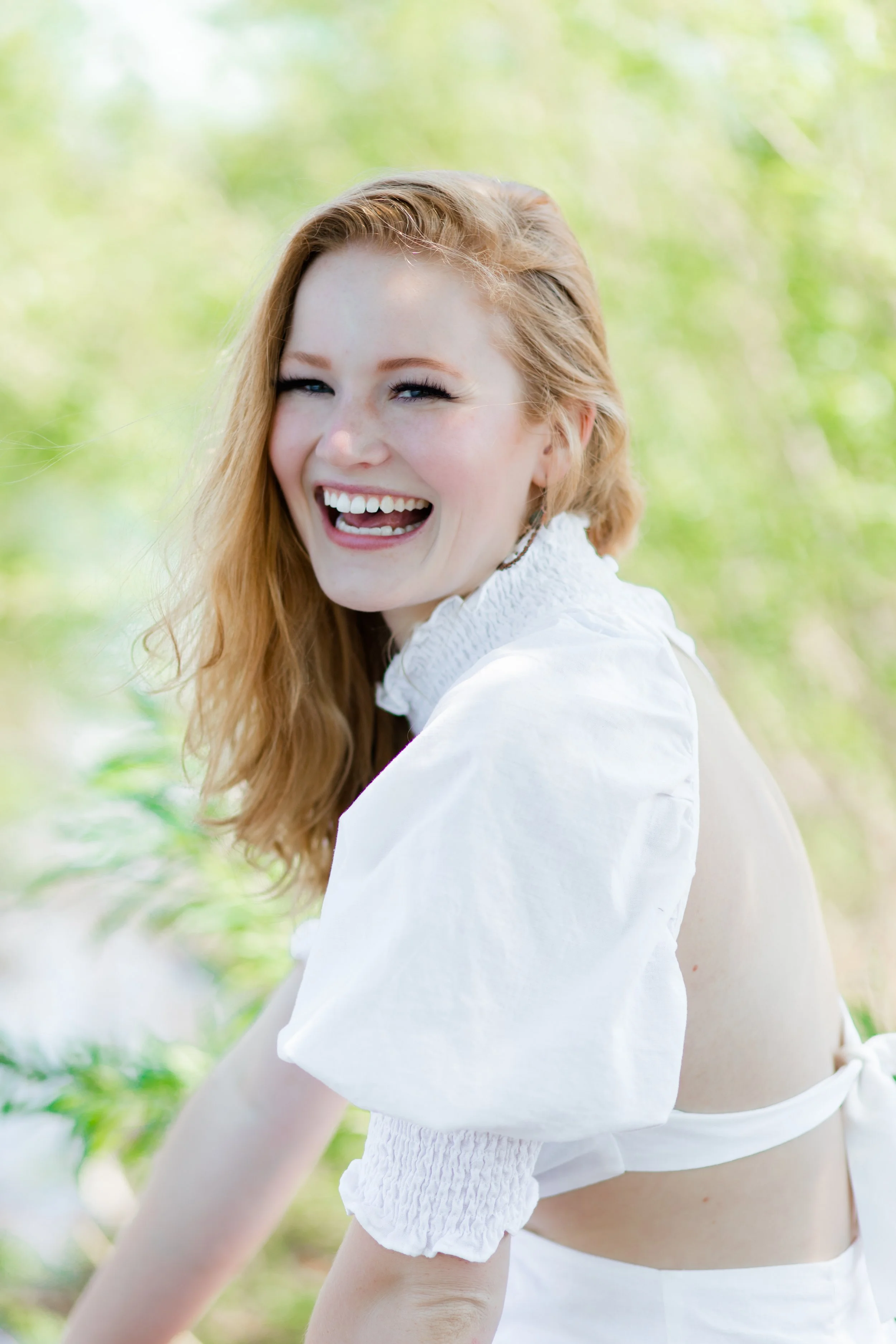 A young woman with long red hair smiling and laughing outdoors, wearing a white blouse, with a blurred green background of trees or plants.