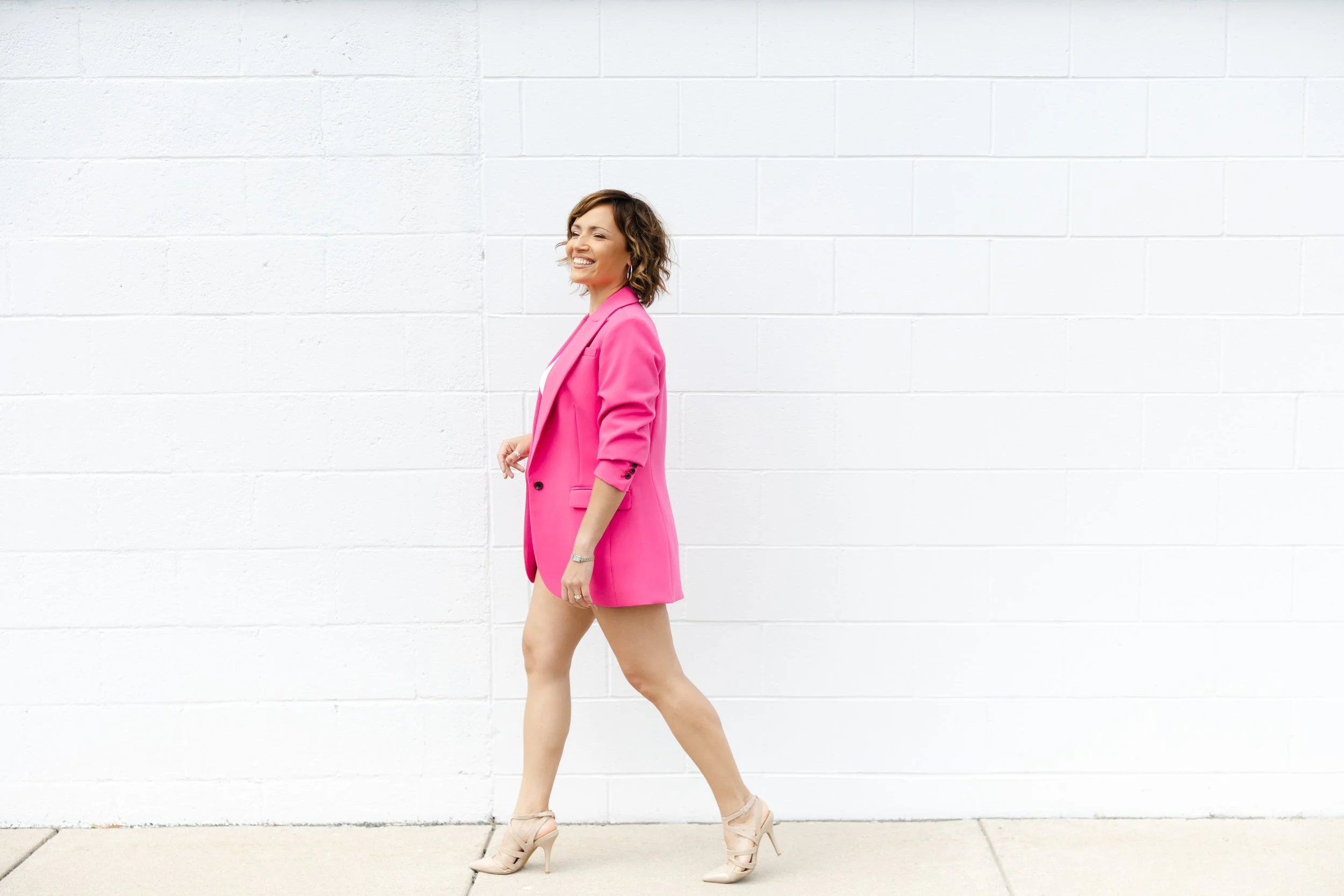 A woman in a pink blazer and matching shorts walking along a white wall, smiling.