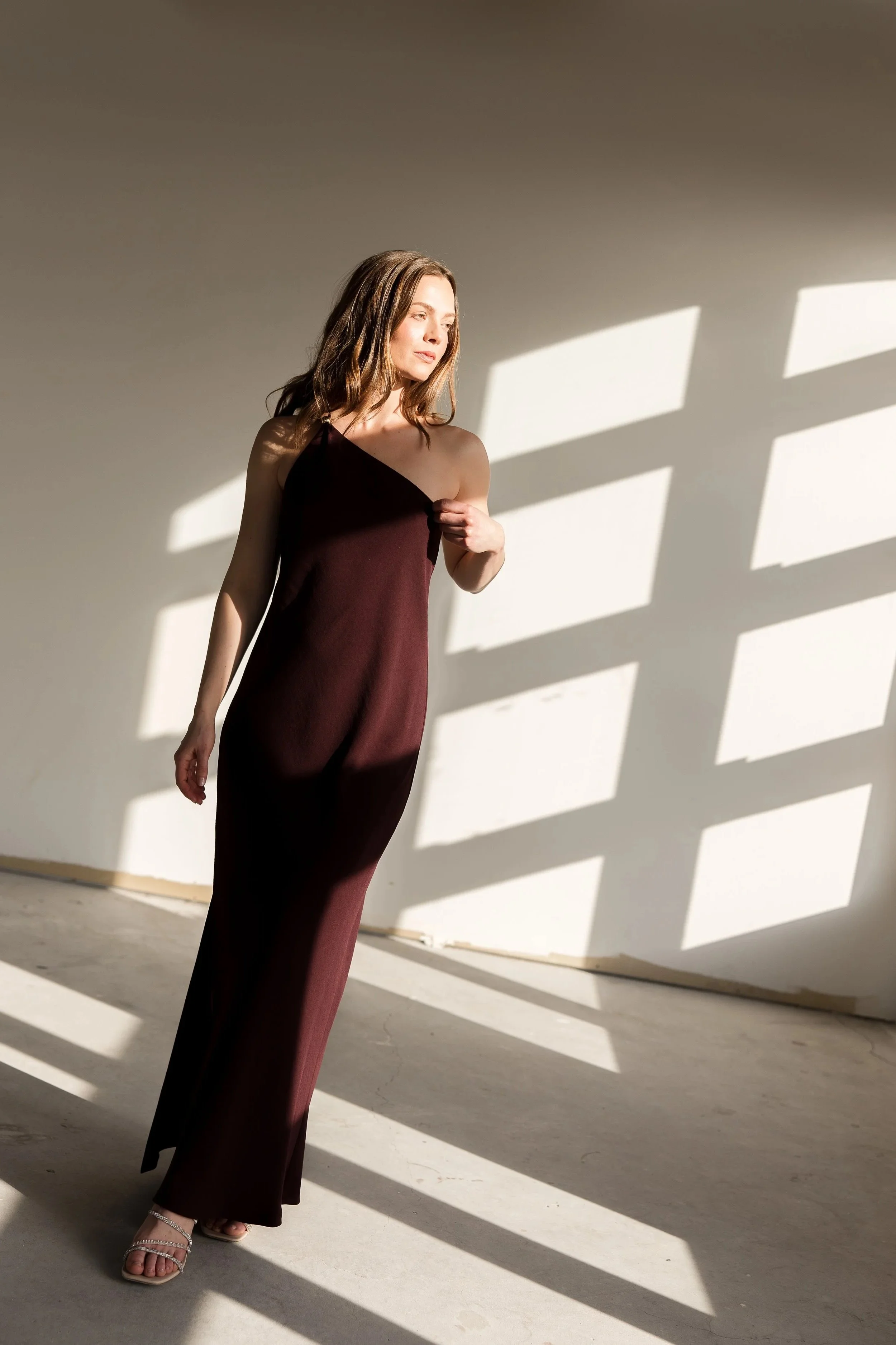 A woman with brown hair wearing a dark red, one-shoulder dress and white high-heeled sandals, standing on a concrete floor with sunlight casting geometric shadows through a window.
