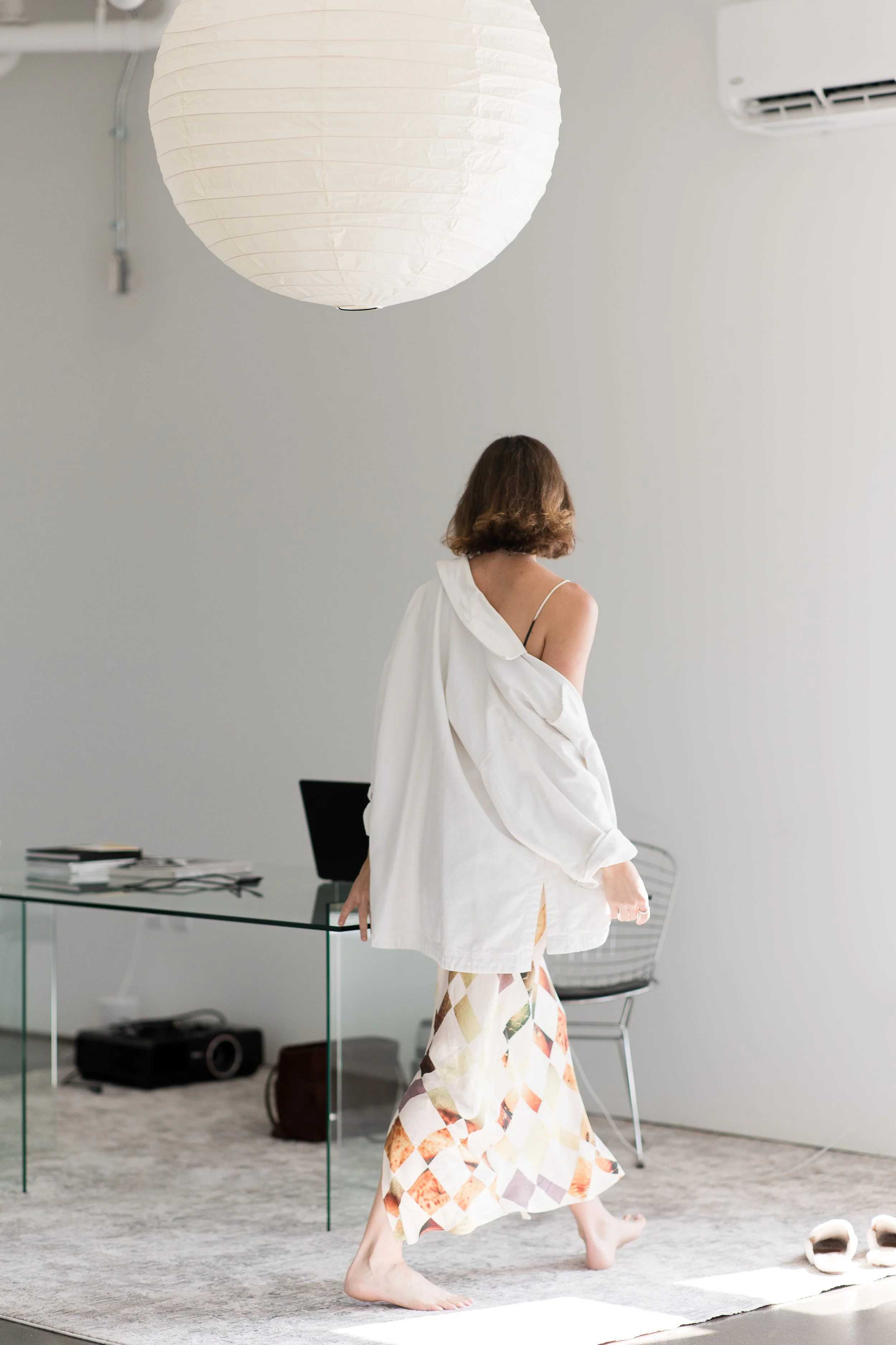 A woman with short brown hair walking barefoot in a bright, minimalistic room with white walls, a glass desk, and a paper lantern ceiling light.
