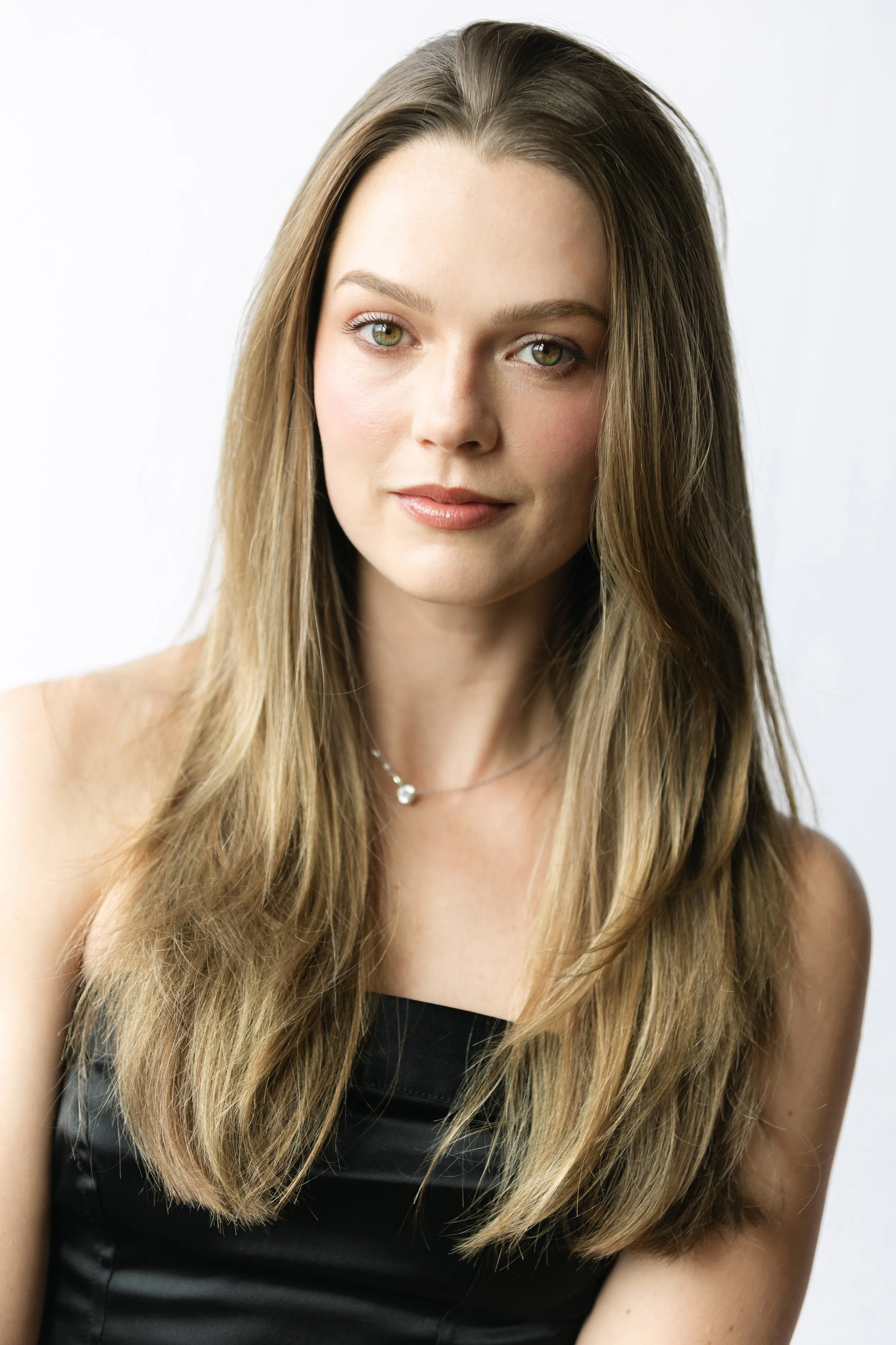 A woman with long, blonde hair, green eyes, and light skin, wearing a black sleeveless top and a delicate necklace, looking into the camera against a plain white background.