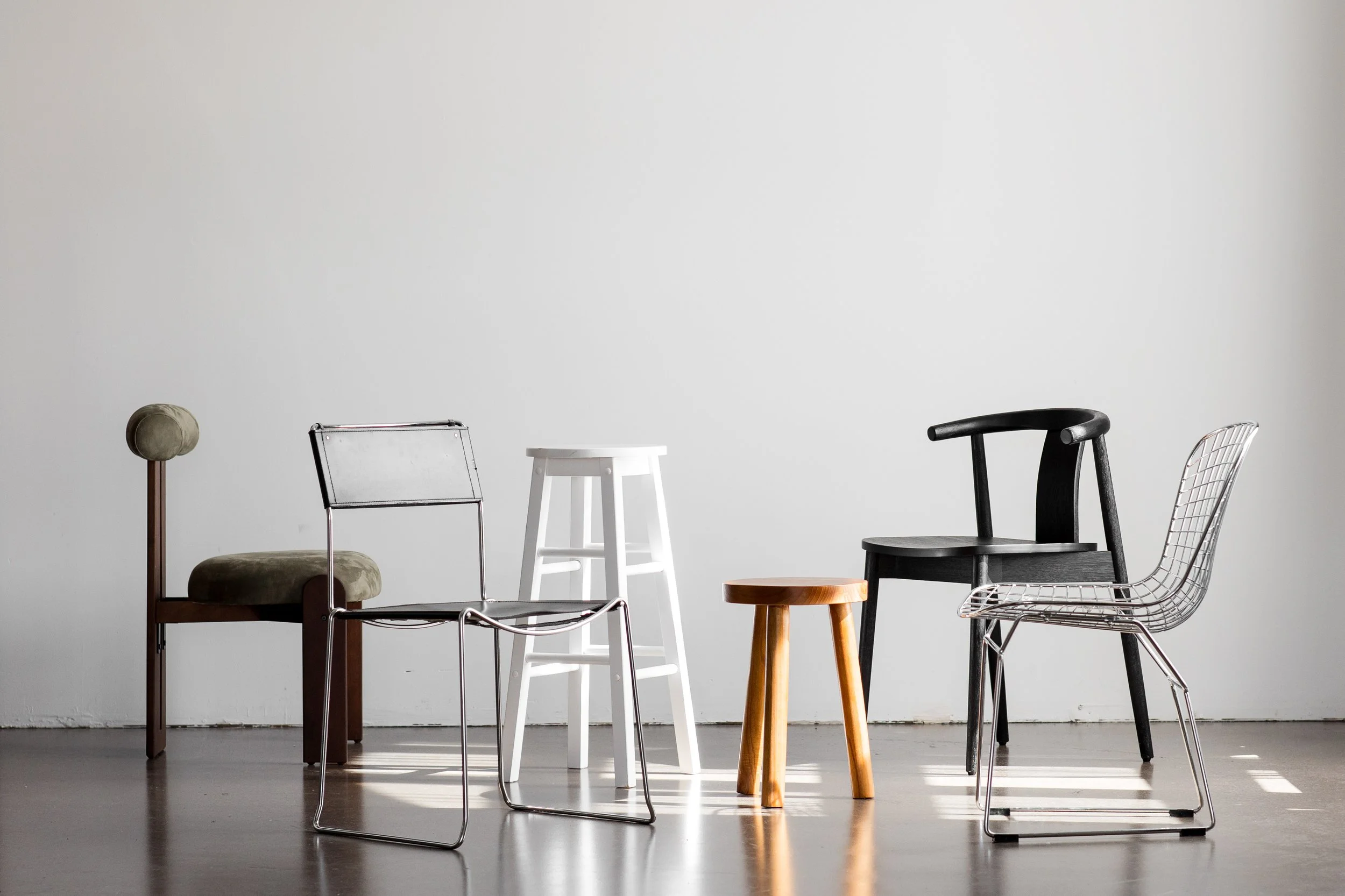 A collection of nine different chairs and stools arranged against a plain white wall, including a beige upholstered chair, a silver metal chair, a white stool, a small wooden stool, a black wooden chair, and a metal wire chair.