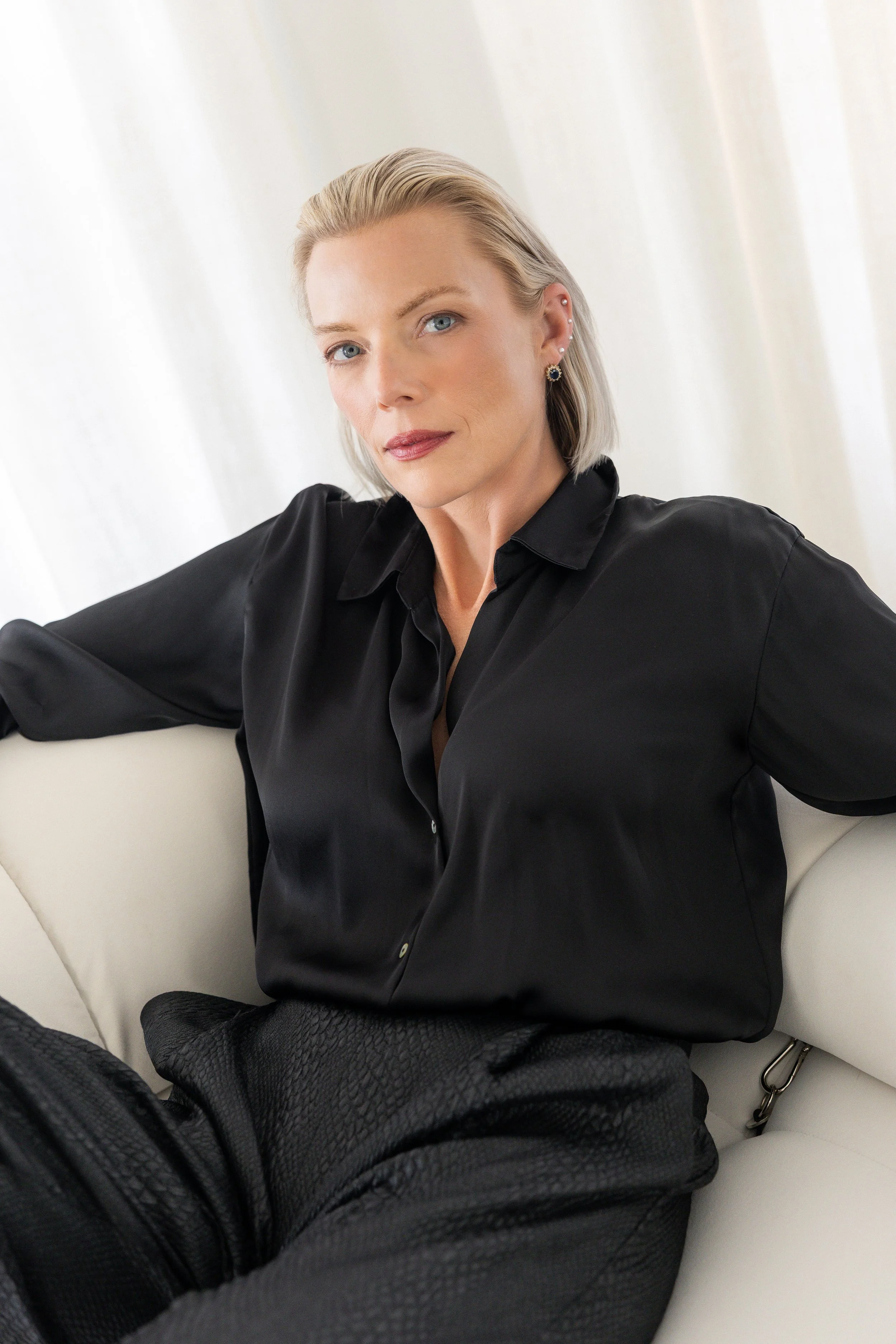 A woman with blonde hair and blue eyes, wearing a black satin shirt, sitting on a white couch, in front of a white curtain.