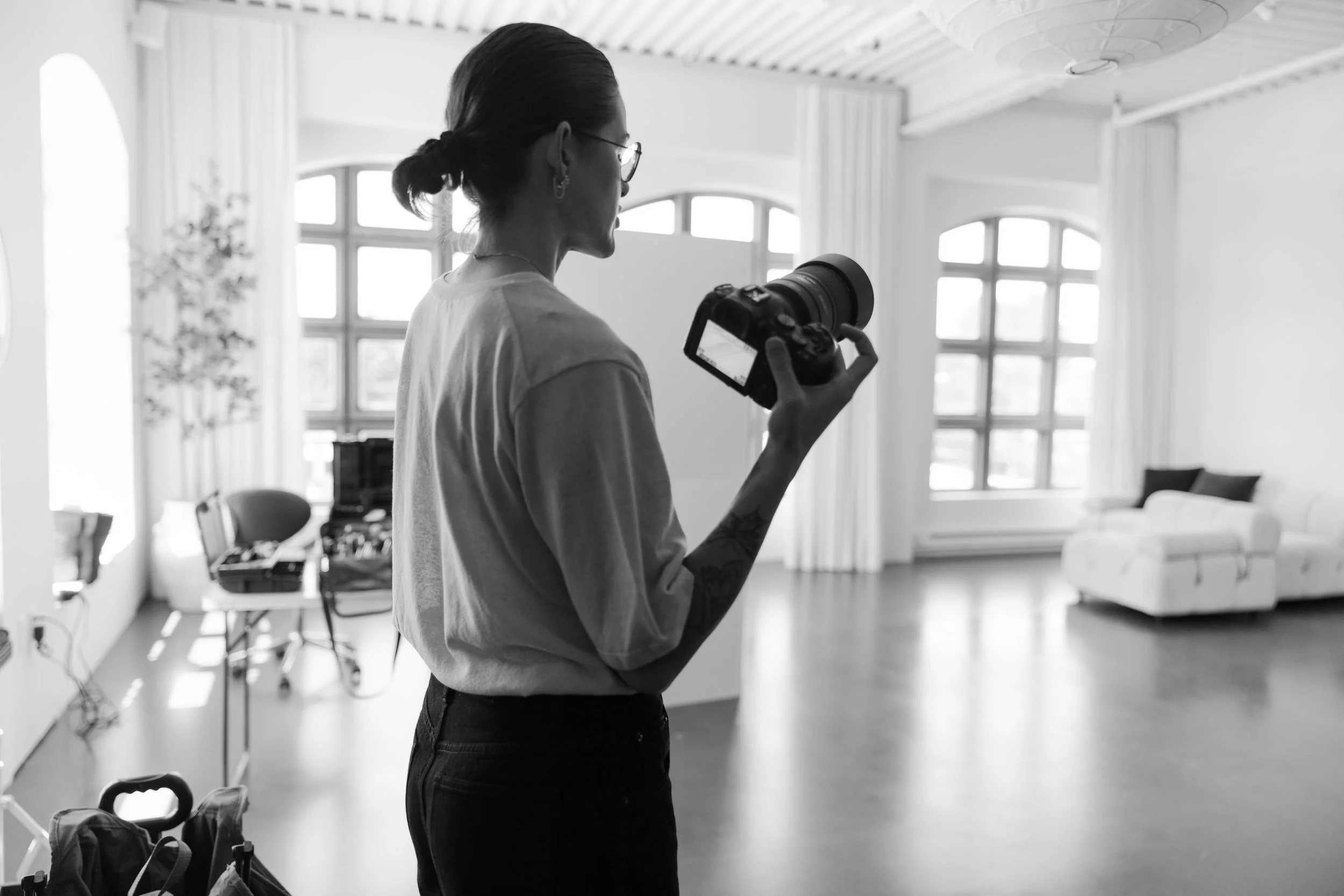 A woman with glasses and a ponytail, holding a camera, standing in a bright, spacious room with large windows, curtains, and a ceiling light.