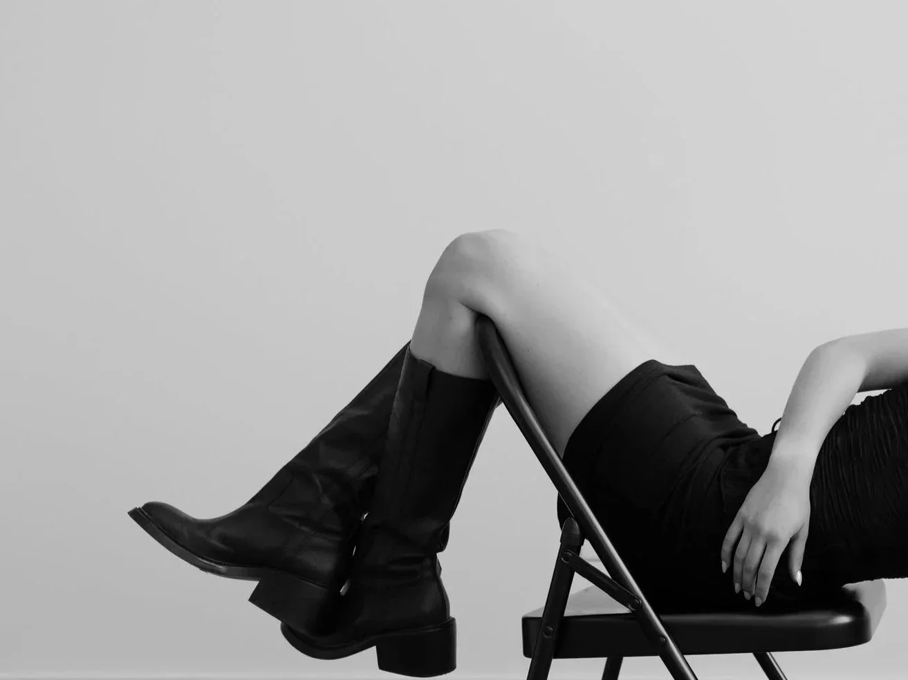 Person sitting on a chair, wearing knee-high boots and a black dress, with legs crossed and one hand resting on their knee, against a plain background.