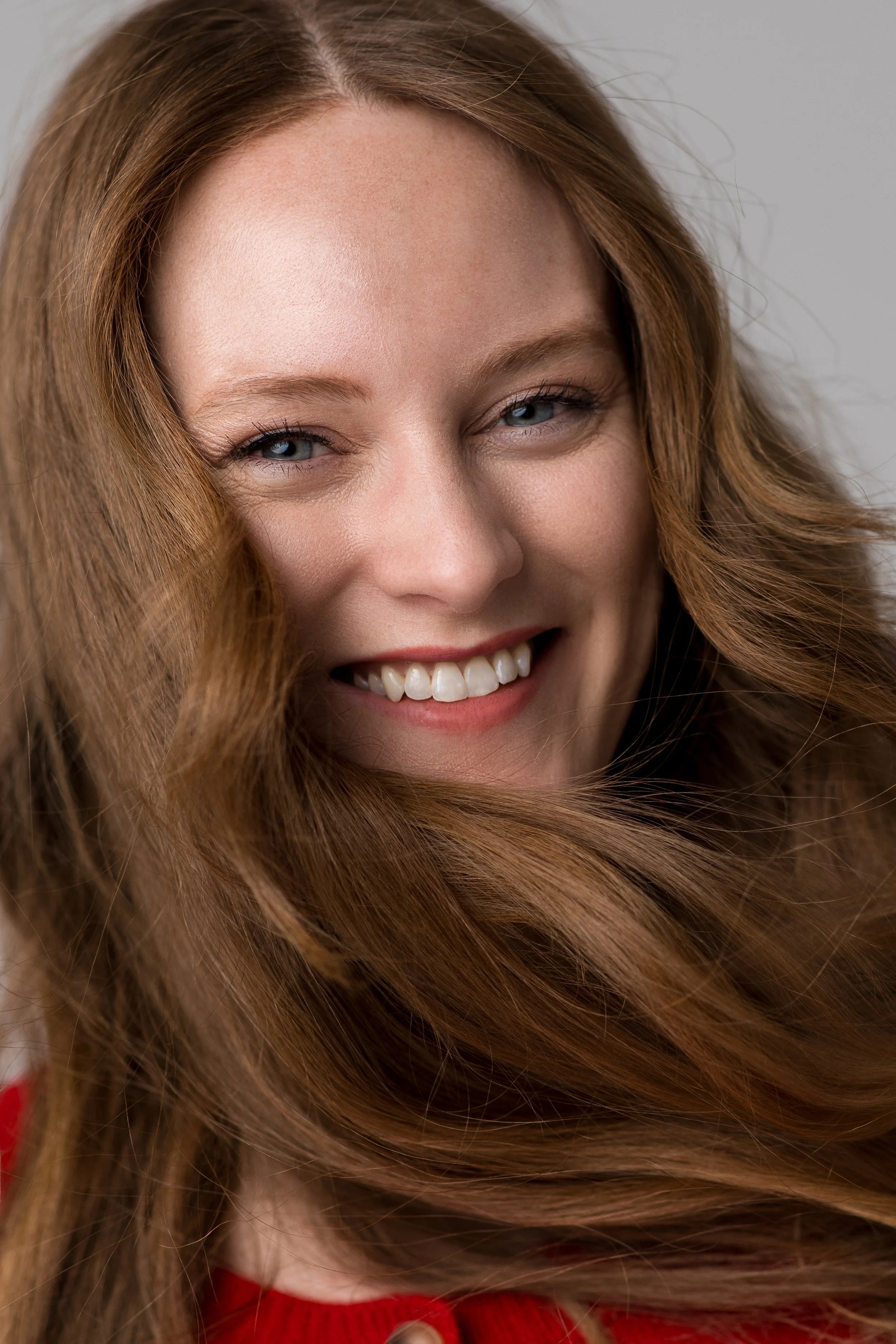 Close-up of a woman with long, wavy red hair, blue eyes, and a big smile, wearing a red top.