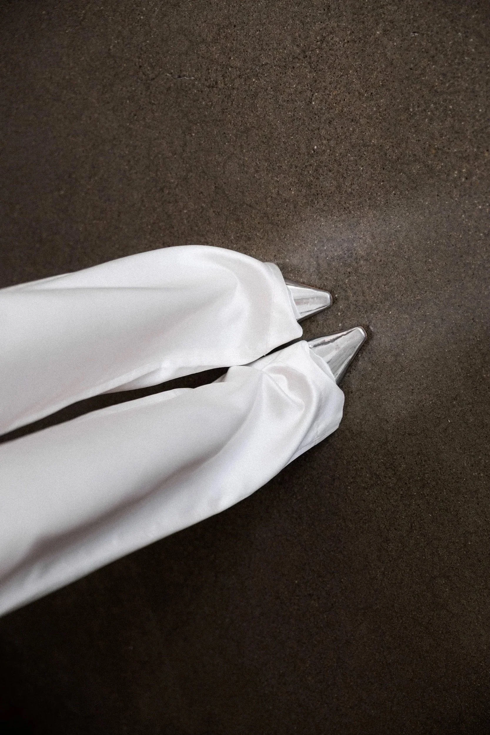 Person wearing white satin dress and silver pointed toe heels standing on dark textured floor.