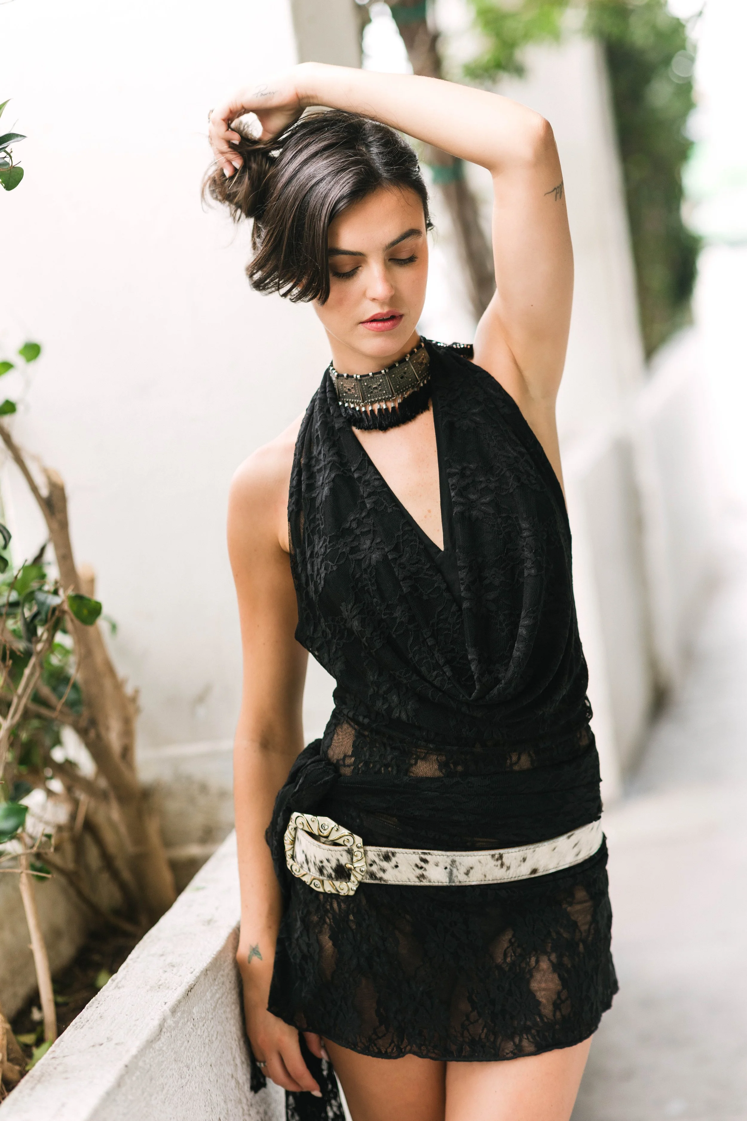 A woman with short dark hair and tattoos on her arms is wearing a black lace dress, a decorative belt, and a statement necklace. She is standing outdoors, leaning against a concrete ledge with greenery and blurred background behind her, and adjusting