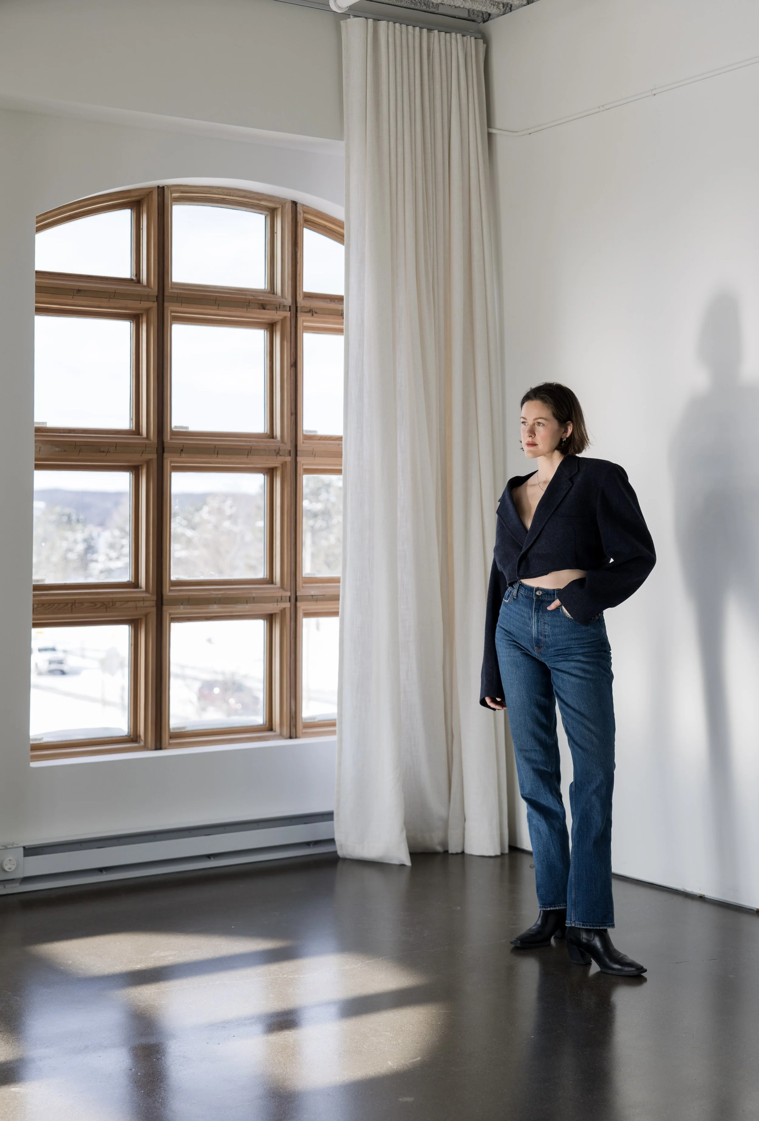 A woman with short dark hair standing indoors in front of a white wall near a large wooden window with snow outside, wearing a dark oversized blazer, high-waisted jeans, and black boots.